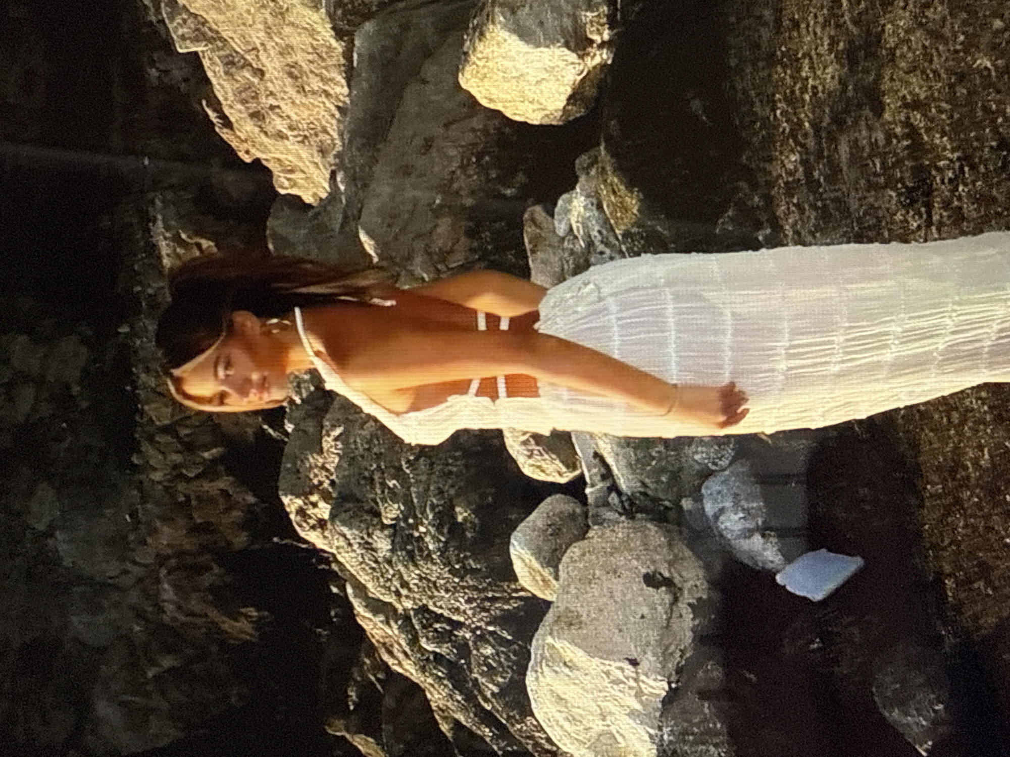 A young woman with her hair in a ponytail, wearing a white halter-neck dress with cut-outs, stands on a rocky terrain. The sun is setting, casting a warm glow on the rocks and the woman. The background is dark and out of focus, with large rocks dominating the scene. The woman is looking over her shoulder to the left, with a slight smile. The location is stated as Găvănoasa, Moldova.