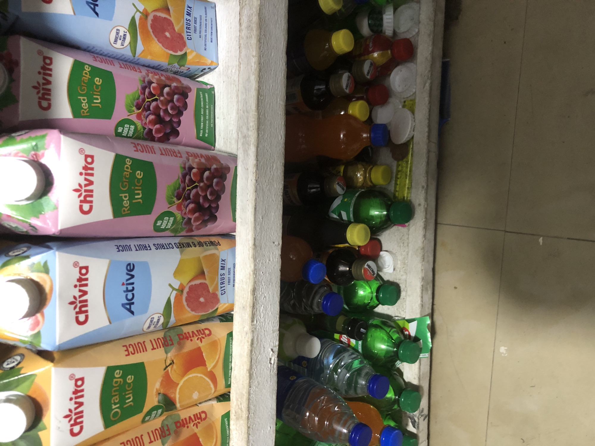 The image displays a close-up view of drinks stacked on shelves. On the top shelf, several Chivita juice cartons are visible, including Orange Juice, Active Citrus Mix, and Red Grape Juice. These cartons are brightly colored with fruit illustrations and branding. Below the top shelf, the second shelf is filled with an assortment of plastic bottles containing various beverages, most of which are green or orange in color. The bottles have colorful caps, predominantly blue, green, and yellow. The background appears to be a wall or shelving unit, with the overall scene suggesting a retail environment or storage area for drinks. The lighting is somewhat dim, casting shadows. There is no indication of a specific location in Ado, Nigeria, within the image itself.