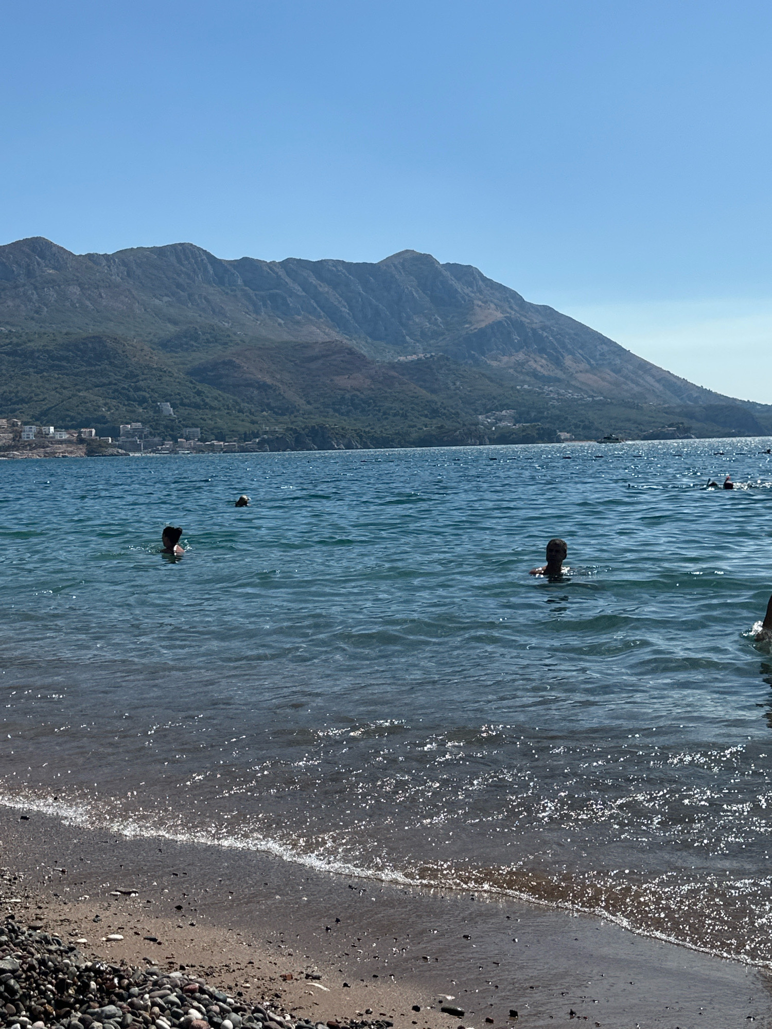 The image depicts a tranquil scene at a beach in Boreti, Montenegro. The foreground features a pebble beach with the gentle lapping of waves. In the water, several individuals are visible enjoying a swim. One person is positioned closer to the shore, with their head and shoulders above the water. Further out, another individual is also swimming. The mid-ground shows more people in the water, some appearing to be snorkeling with visible masks and snorkels. The background is dominated by a large, majestic mountain range, its slopes covered in lush greenery and dotted with buildings, indicating a coastal town. The sky is a clear, bright blue, suggesting a sunny day. The overall atmosphere is peaceful and idyllic, characteristic of a summer day at the beach.
