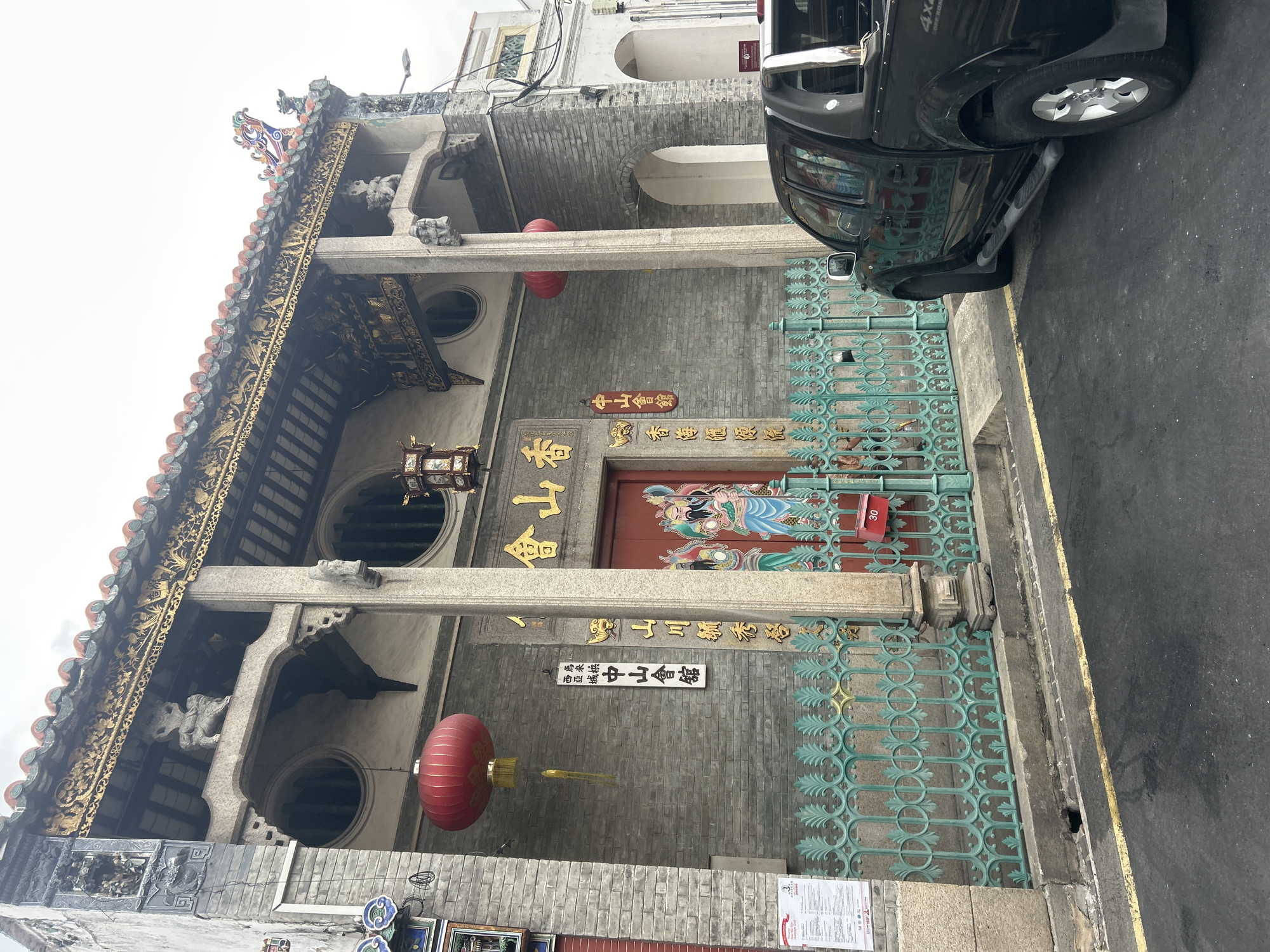 The image depicts the exterior of the Yap Temple, also known as the Kong Tat Kong Temple, located on the busy Carnarvon Street in the heart of George Town, Penang, Malaysia. The temple facade is characterized by its traditional Chinese architectural style, featuring intricate carvings, ornate eaves, and prominent columns. Red Chinese lanterns hang in front of the building, adding a festive touch. A turquoise wrought-iron fence encloses the temple grounds, with a black pickup truck parked on the street in the foreground, partially obscuring the view of the entrance. The street itself is paved with asphalt and shows signs of wear. The overall scene captures a blend of traditional heritage and the urban environment of George Town.