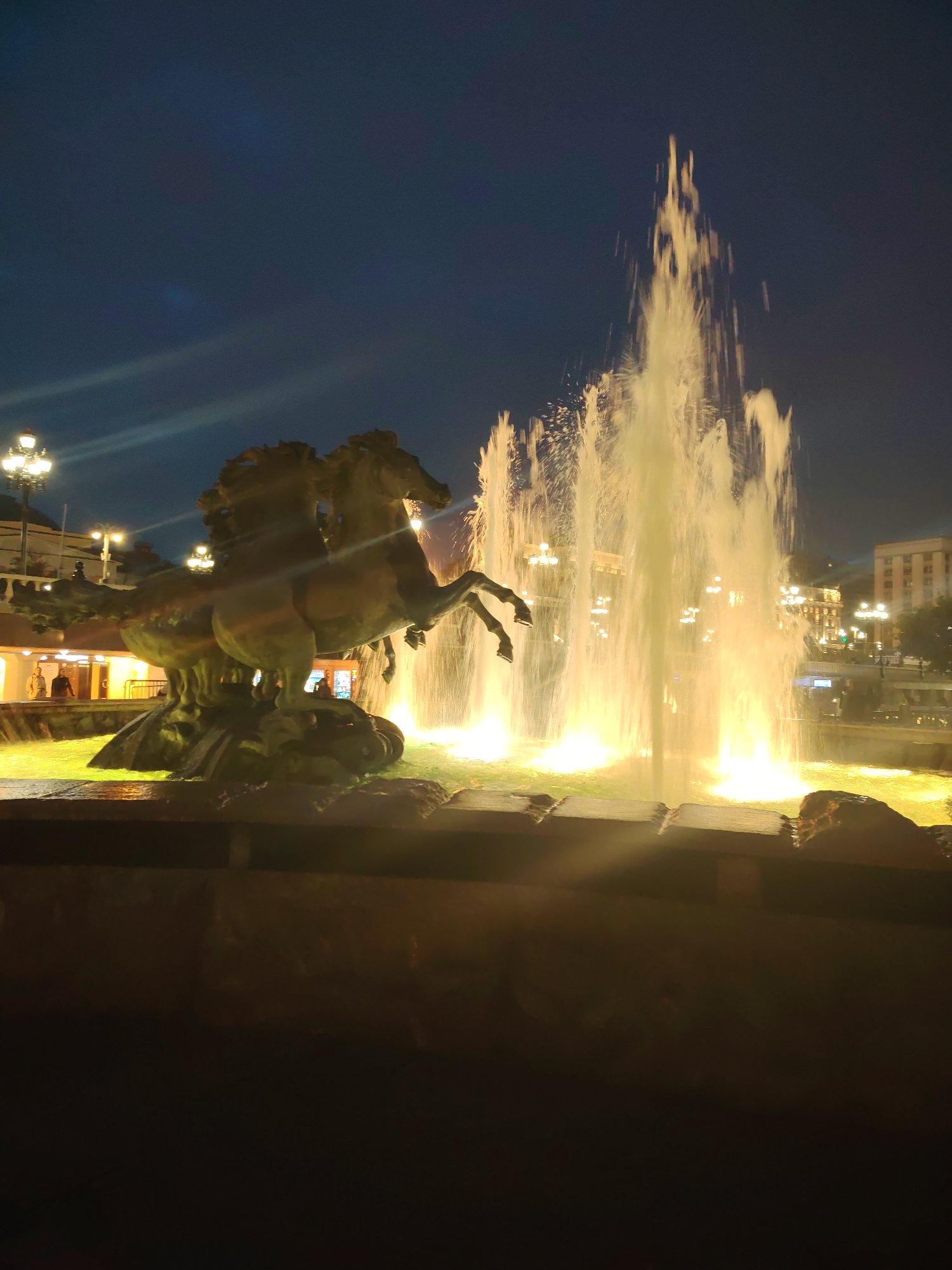 A nighttime, medium-shot photograph captures a monumental fountain at the center of the frame. The fountain features a sculpture of four horses in mid-gallop, their bodies rendered in a bronze-like material, positioned on a rocky base. The horses are dynamic, with their heads raised and legs extended, suggesting movement and power. A central, powerful jet of water erupts from behind the horses, illuminated by warm, golden lights that create a dramatic glow. The water cascades down, forming a misty veil around the sculpture and pooling in the basin below, which is also lit with the same golden hue, giving the water a vibrant, almost molten appearance.
In the background, a cityscape unfolds under a dark, deep blue night sky. The city is dotted with numerous streetlights, their warm illumination creating a soft glow that outlines buildings and lampposts. These lights are mostly of a traditional design, with multiple bulbs on ornate poles. Further in the distance, the lights of larger buildings suggest a bustling urban environment.
On the left side of the image, a few people are visible near a low wall, interacting with the fountain area. They appear to be casually observing the spectacle. The overall atmosphere is one of grandeur and nocturnal beauty, highlighting the interplay of light, water, and sculpture. The scene is set outdoors, in a public urban space that has been transformed by nighttime illumination.