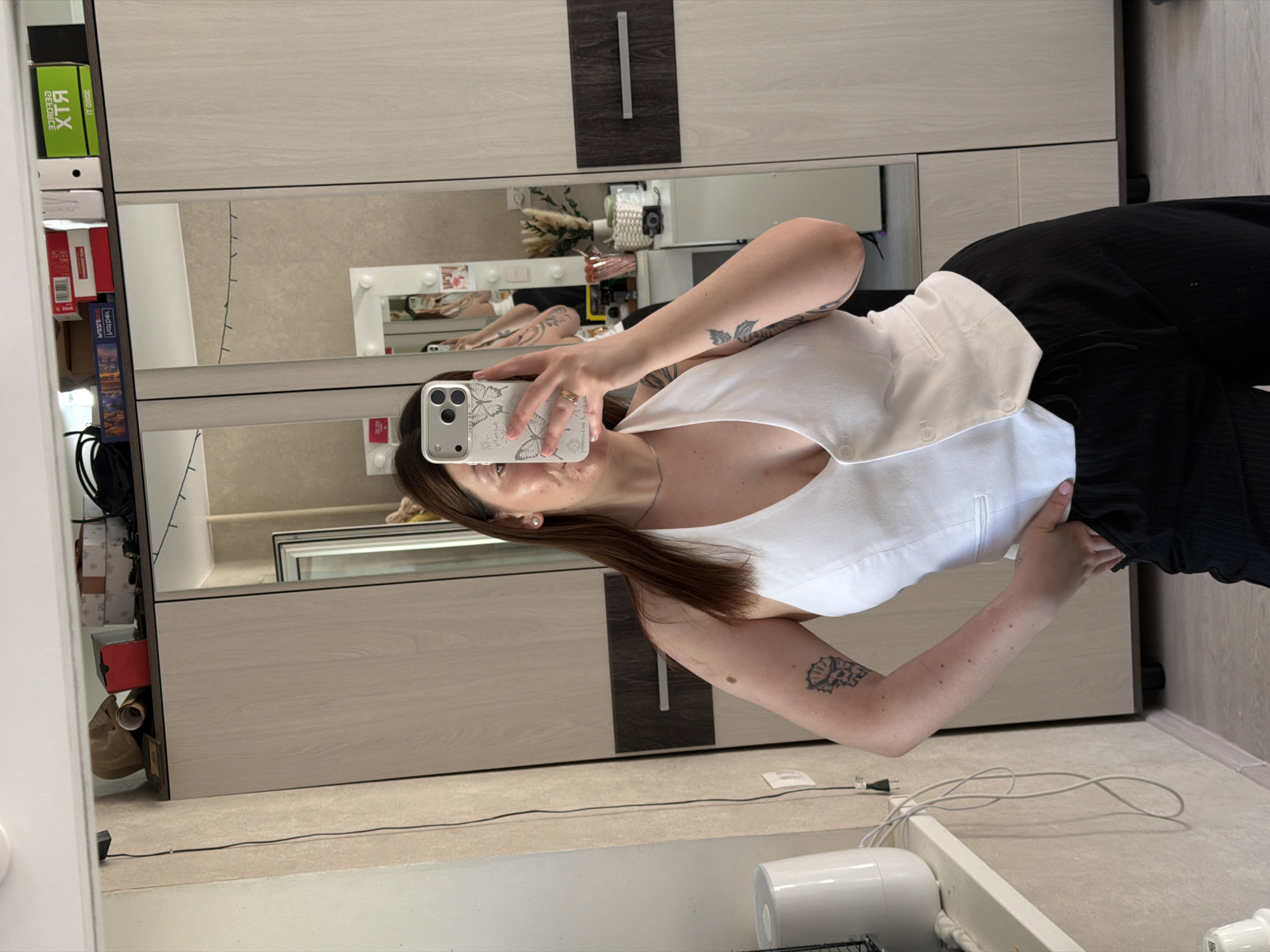 The image shows a woman taking a selfie in front of a mirror. She is wearing a white top and dark pants. The background includes a wardrobe and other items on the floor, such as a heater. There is a reflection of the woman and objects behind her in the mirror. The location is implied to be in Chaikovsky, Russia, based on the user's prompt. However, there is no readily available visual evidence to confirm that location.