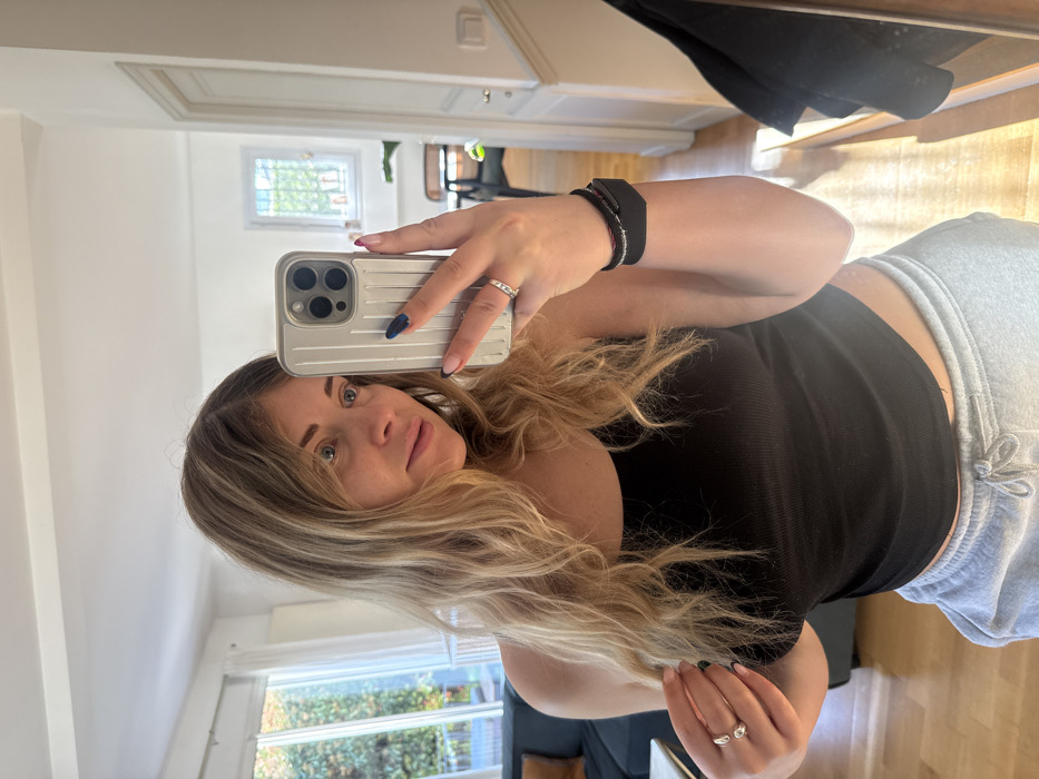 A female is depicted in a mirror selfie within a residential interior in Rueil-Malmaison, France. She possesses long, wavy blonde hair and blue eyes, and is attired in a black ribbed tank top and light grey drawstring sweatpants. Her right hand holds a silver-colored smartphone featuring a triple-lens camera array and a ribbed protective case, positioned to capture her reflection. On her right wrist, a black fitness tracker and a slender black bracelet are visible. Her right ring finger displays a silver ring with embedded stones, and her nails exhibit dark blue/black polish with light pink tips. Her left hand is raised, touching her hair, revealing two silver rings on her left ring finger (one a plain band, the other an open design) and nails with dark-colored tips.
The background, observed through the mirror, consists of white walls and light wooden flooring. To the left, a large glass door or window provides a view of dense green foliage outside, and a dark blue sofa is partially visible indoors. To the right, a white-framed window with exterior security bars shows a bright sky and distant trees. A white doorway leads into an adjacent room where a dark chair and a hanging dark garment are present. The scene is illuminated by natural light.