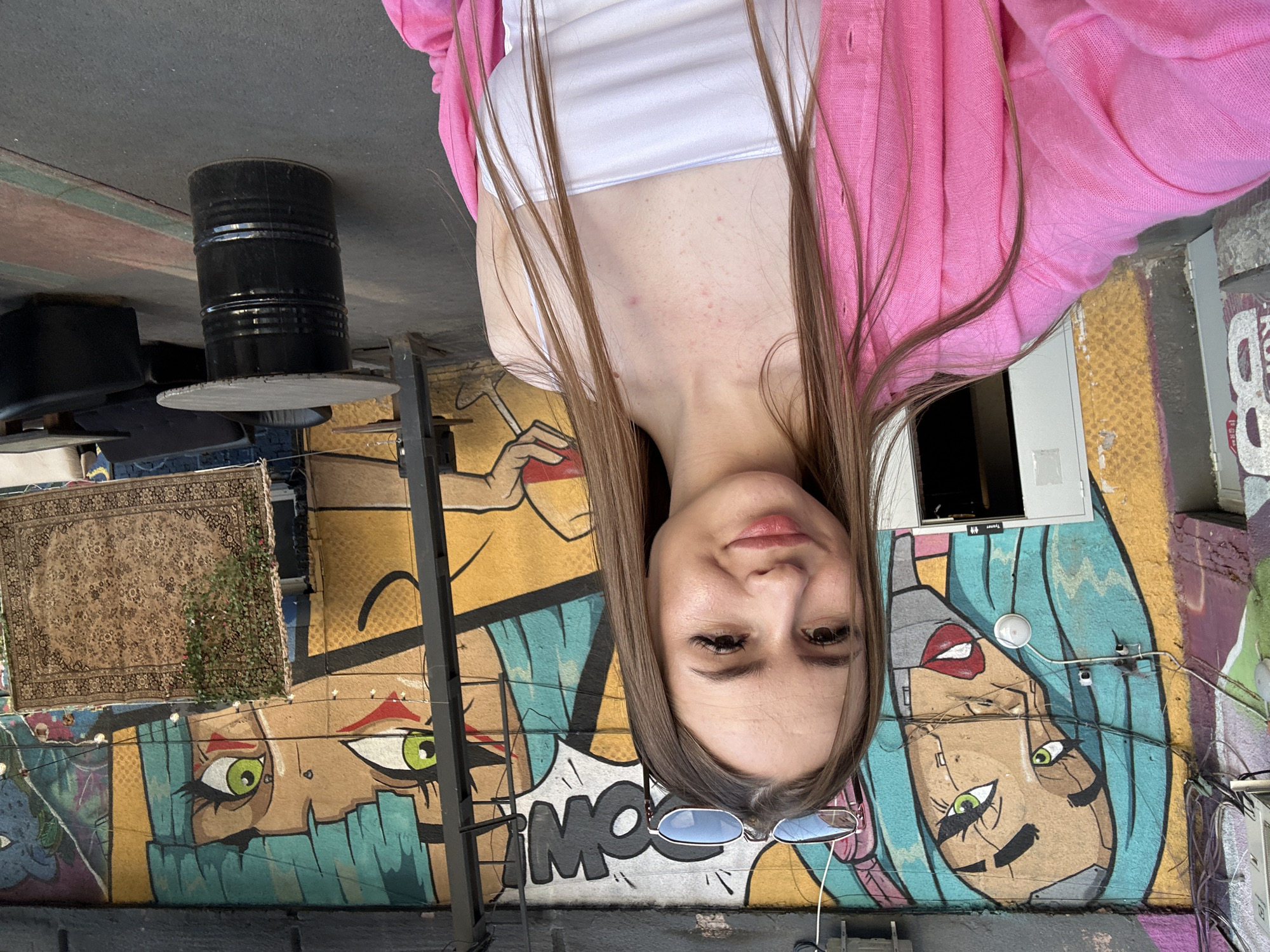An inverted close-up portrait features a young woman positioned outdoors against a backdrop of vibrant street art. The woman has long, straight light brown hair flowing around her face and shoulders, looking directly at the viewer with a calm, slightly smiling expression. She is wearing a white crop top layered under a bright pink, open-front knitted cardigan or jacket. A pair of blue-lensed sunglasses with light frames rests on top of her head.
The background is dominated by extensive and colorful graffiti murals. These murals depict stylized faces with bold features, including large green and brown eyes and hair in shades of blue and black. One prominent section of the mural shows a character with partially robotic or cyborg-like elements. Speech bubbles are integrated into the art, one of which visibly contains the partial word "BOOM!". Other textual elements like "TYRANTA" and "88" are also visible within the graffiti.
To the left of the woman, a black metal barrel topped with a circular surface functions as a table base. Above this, a large, intricately patterned rug is hung vertically on a wall adorned with graffiti. The setting conveys an artistic urban environment, possibly a trendy cafe or an art district, during daylight hours. The image captures an informal moment, highlighting the woman's relaxed presence amidst the dynamic and eclectic surroundings.