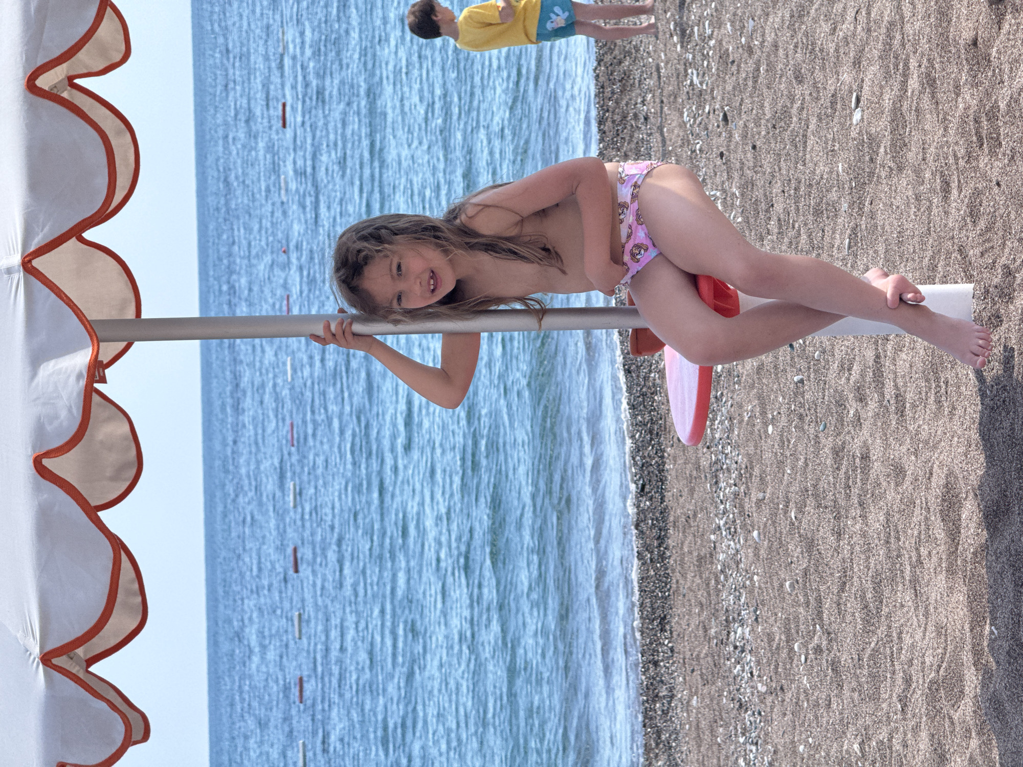 The image shows a young girl at the beach. The girl is leaning on a silver pole with a white beach umbrella attached to it. She is wearing pink swim bottoms with a cartoon print and a red boogie board is positioned next to her. Her head is to the left and her legs are extended on the pole. The ocean is behind her, and in the background, there is another person standing on the shore. The beach is made of sand and small pebbles. The location is Boreti, Montenegro.