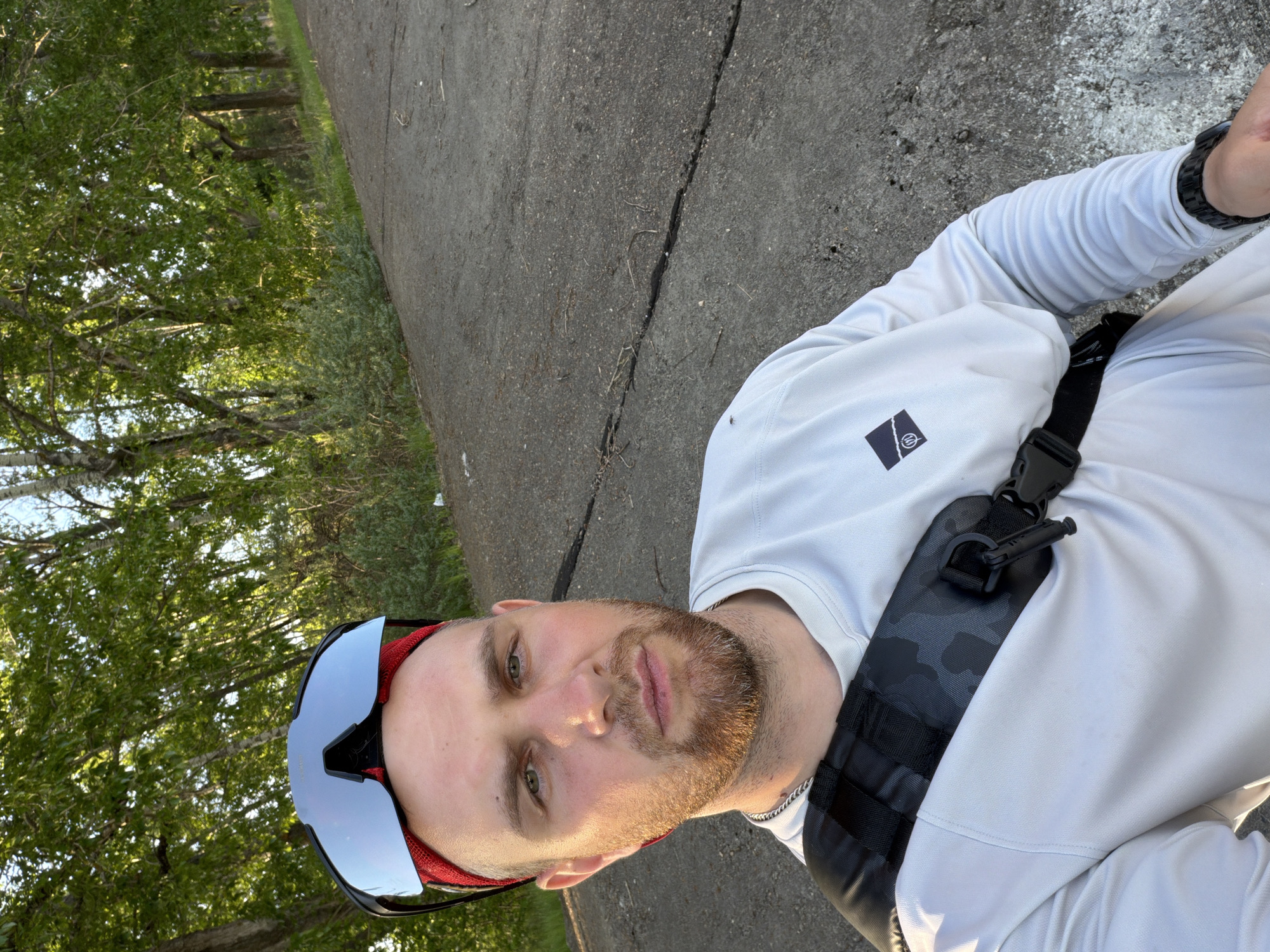 A selfie of a light-skinned man with a short beard, wearing a white long-sleeved shirt, sunglasses, and a black chest strap, likely taken on a paved path in a park or green area. Trees are visible in the background. The location is possibly in the city of Chaikovsky, Russia, but this cannot be verified from the image alone.