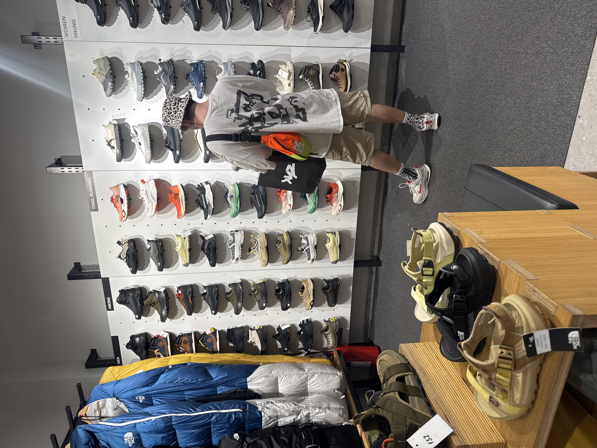 The image shows the interior of a retail store, likely a North Face or similar outdoor gear store, potentially in Belgrade, Serbia. A person wearing a white graphic t-shirt, beige shorts, and patterned socks with running shoes is standing in front of a wall display of shoes. The person also has a leopard print hat, a black shoulder bag with a white logo and an orange fanny pack. The wall displays rows of shoes categorized as 'Women's Hiking' and 'Trail Running' shoes. Below the shoe display, there are stacks of blue and silver sleeping bags and black backpacks. To the right of the person, there is a wooden display unit containing various sandals and footwear. The floor is a combination of gray carpet and concrete.