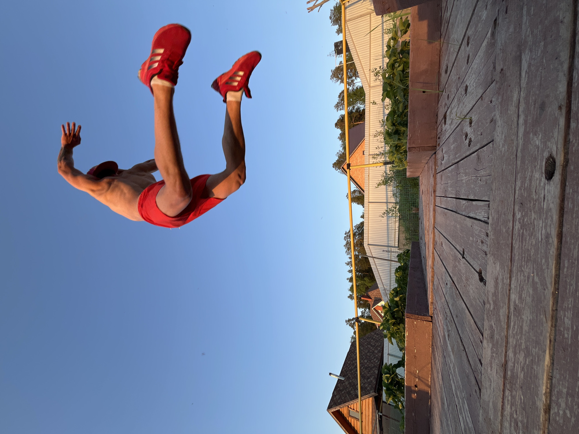 A man is captured mid-air against a clear blue sky. He is wearing red shoes, red shorts, and a red hat. His body is turned upside down, with his arms and legs extended. The backdrop includes a wooden structure, possibly a deck, and glimpses of residential buildings with varying rooflines and light-colored siding. A yellow gas pipe runs alongside a building. The image appears to be taken from a low angle.
The location is likely Chaikovsky, Russia, based on the user's prompt.