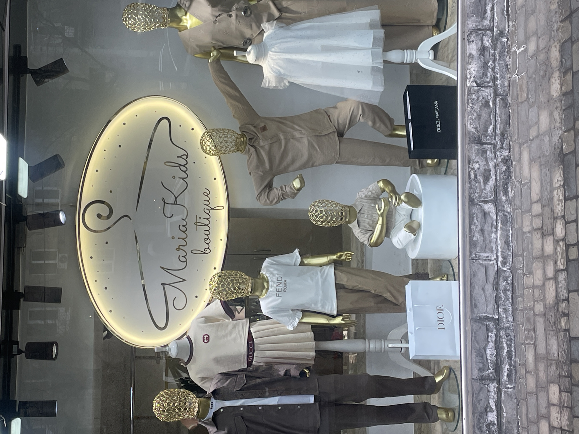 The image displays a storefront window of "Maria Kids boutique" in Yerevan, Armenia. Six golden-skinned mannequins, all featuring abstract, mesh-like golden heads, are arranged within the window.
From left to right:
1. A standing mannequin wears a dark brown long-sleeved shirt unbuttoned over a white t-shirt with "BOSS" visible on the collar, paired with dark brown pants.
2. A standing mannequin, positioned on a white pedestal, wears a cream-colored short-sleeved crop top with a red circular logo (Gucci) and a matching pleated mini-skirt with "GUCCI" visible on the waistband.
3. A standing mannequin wears a white short-sleeved t-shirt with "FENDI ROMA" printed in black, and light brown pants.
4. A small, seated mannequin rests on a white cylindrical pedestal, wearing a light brown short-sleeved polo shirt and white pants, holding a golden scoop-like object.
5. A standing mannequin in a dynamic pose, leaning right with an extended left arm, wears a light brown long-sleeved button-up shirt and matching pants.
6. A standing mannequin wears a light brown sleeveless vest with a subtle embroidered pattern and matching wide-leg pants. To its left, a white child's dress is displayed on a white stand.
An illuminated oval sign at the top center of the display reads "Maria Kids boutique" with a hanger graphic and small light dots. Black track spotlights are mounted above, illuminating the scene. On the display floor, a white "DIOR" shopping bag is visible on the left, and a black "DOLCE&GABBANA" box is on the right. Reflections of an urban environment, including buildings and trees, are visible in the glass. The exterior base of the window features a textured wall of gray stone blocks and bricks.