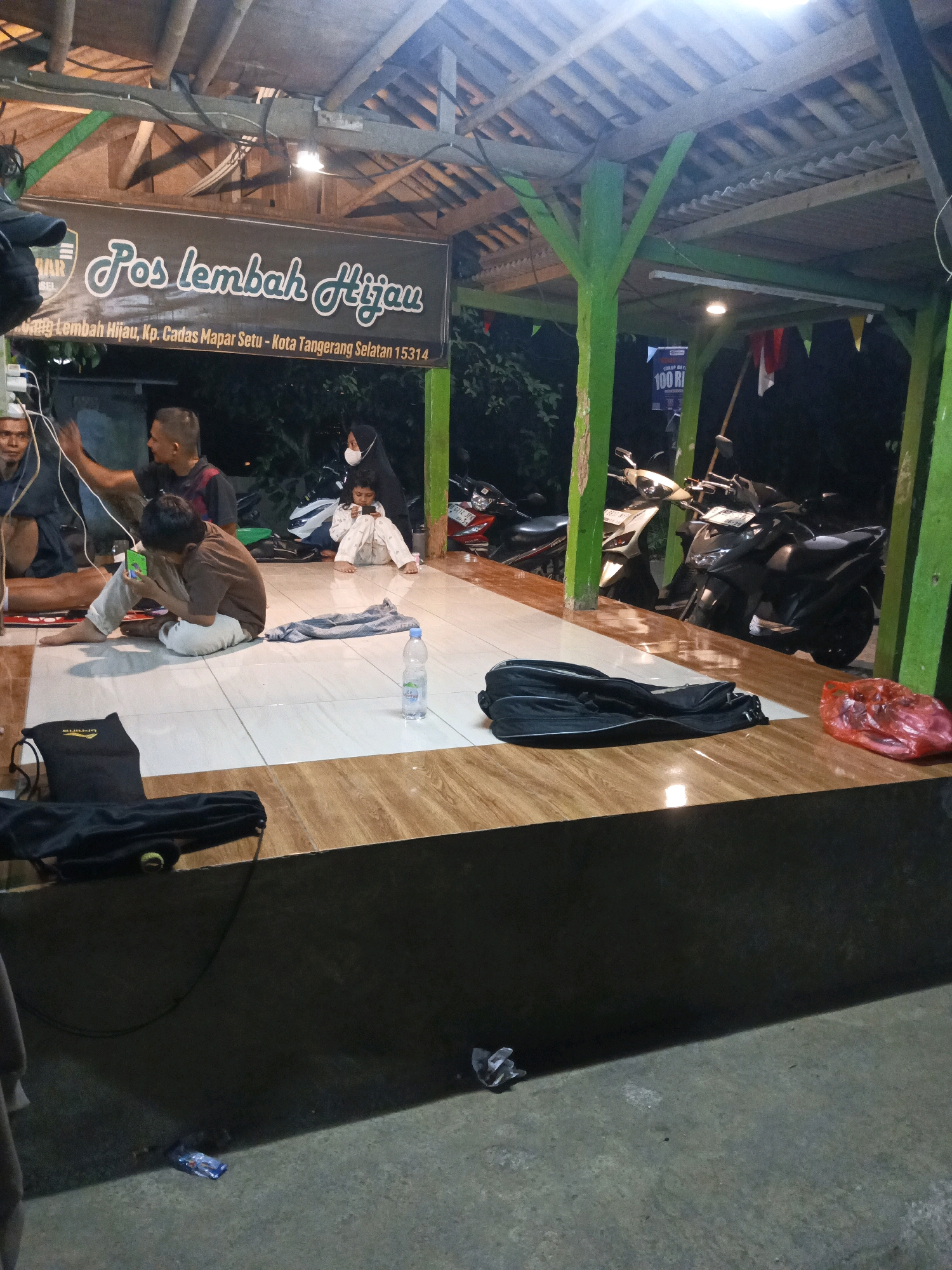 The image shows an outdoor seating area at "Pos Lembah Hijau" located at Kp. Cadas Mapar Setu in South Tangerang, Indonesia. A group of people are seated on a tiled floor under a wooden roof supported by green pillars. In the foreground, a black bag lies on a wooden bench. A red plastic bag is situated next to it. To the left, another black bag is visible on the floor.
A boy in a brown shirt and white pants is sitting and looking at a mobile phone. Next to him, a woman wearing a black hijab and mask is holding a child who is also looking at a phone. To the left, two men are seated. One man is wearing a dark shirt and the other is wearing a dark t-shirt with a red accent. One of the men appears to be gesturing.
In the background, several motorcycles are parked. A sign above the seating area reads "Pos Lembah Hijau" and provides the address. Another sign in the background shows "100 RI". A bottle of water sits on the tiled floor. The overall lighting suggests it is evening or nighttime.