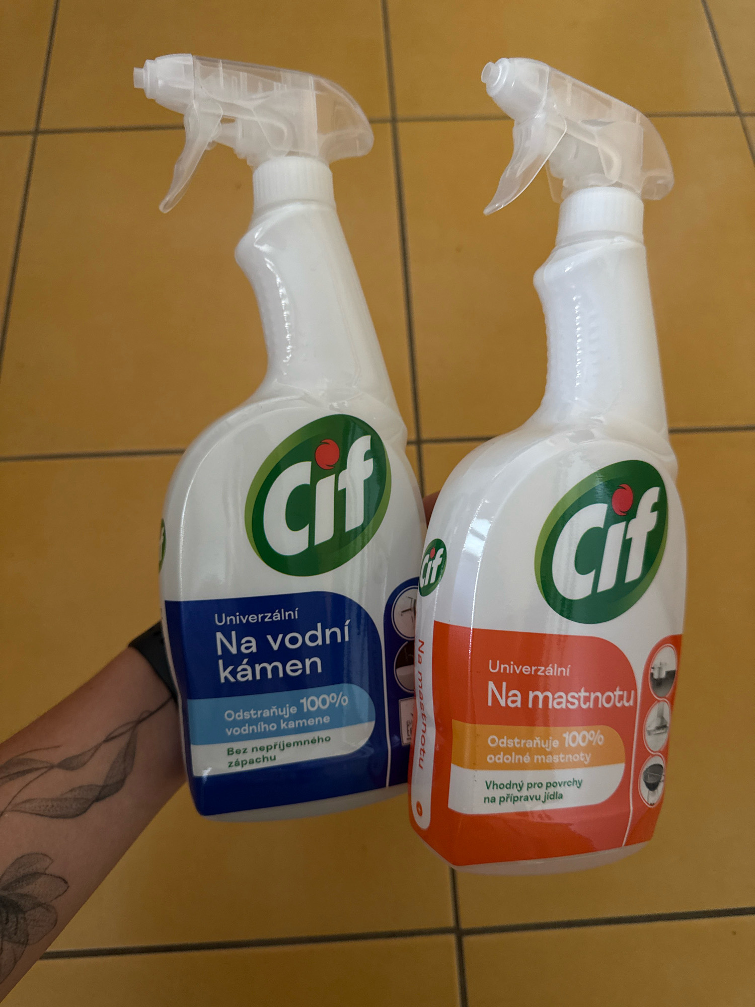 The image shows two Cif brand spray bottles of cleaning products against a yellowish tiled background. The bottle on the left is labeled "Univerzální Na vodní kámen" (Universal for limescale) and the bottle on the right is labeled "Univerzální Na mastnotu" (Universal for grease). The bottles are standing upright. A person's arm with a tattoo is holding the left bottle. The background appears to be floor tiles. The location is not specified in the image.