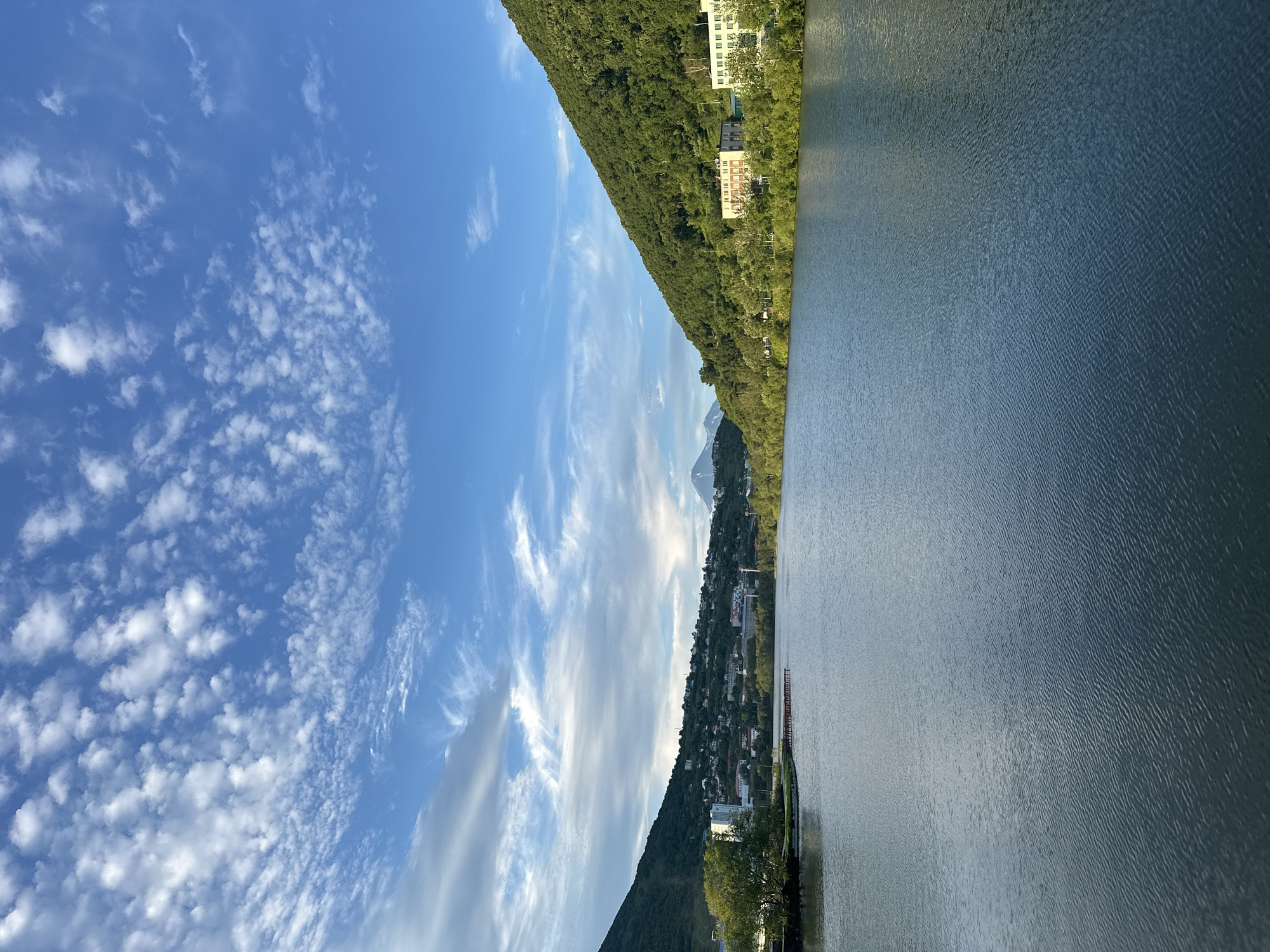 The image shows a body of water with a town built on a hill in the background. The water is calm with small ripples. On the left side of the water, there is a small park area with a tree and a pier. The town on the hill has many houses and buildings. On the right side, there is a lush green hill with several buildings visible. In the far distance, there are mountains. The sky is blue with scattered white clouds. The scene appears to be a scenic view of Petropavlovsk-Kamchatsky, Russia, likely overlooking the Avacha Bay or a similar body of water within the city.