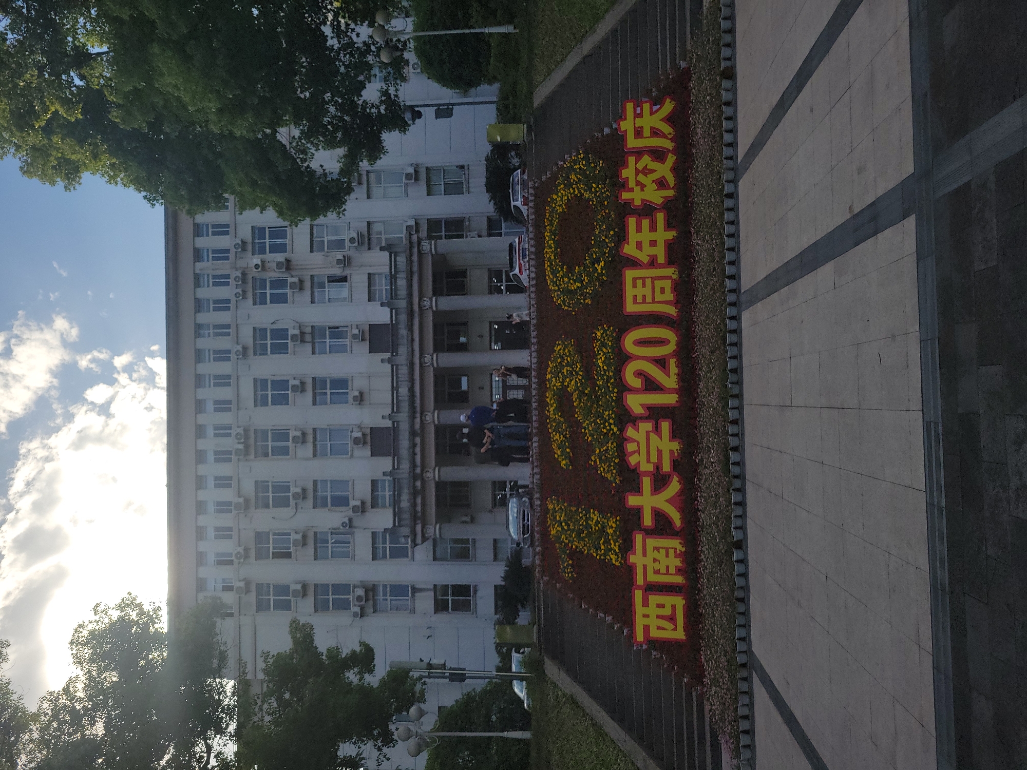 The image shows a building with a sign celebrating the 120th anniversary of Southwest University, in Beibei, China. The building is white, several stories high, and has many windows. The sign is made of red and yellow flowers, arranged to form Chinese characters. People are standing in front of the building. Cars are parked nearby. The setting appears to be a university campus.