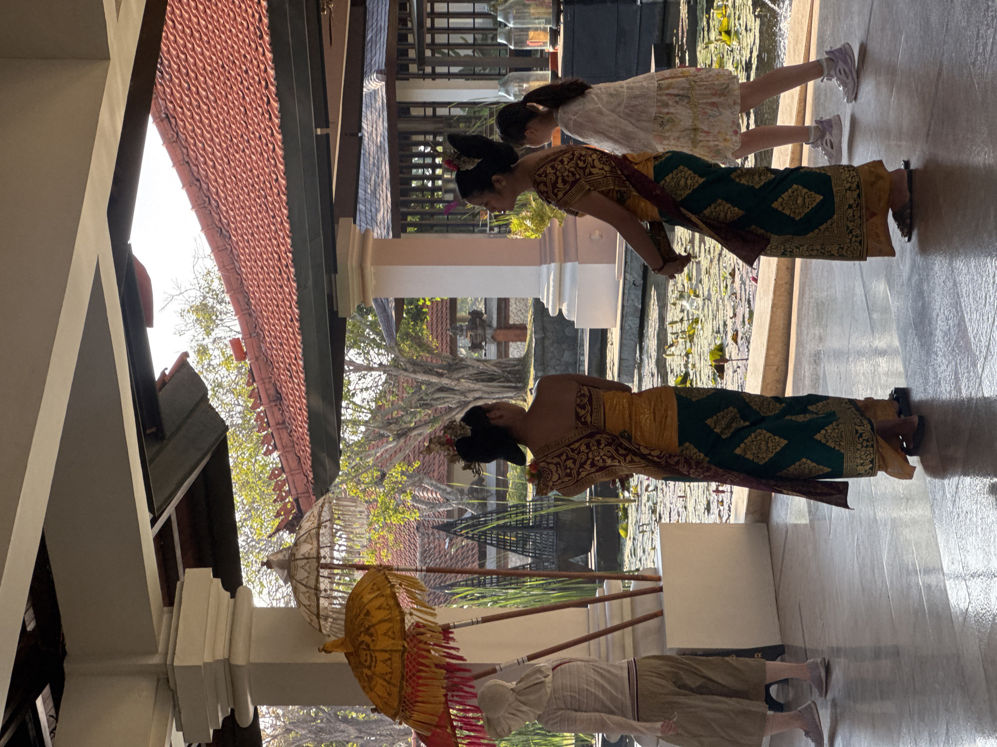 In Kuta Selatan, Indonesia, two women in traditional Balinese attire, a golden-yellow top with a maroon and gold patterned sash and a dark green patterned sarong, stand facing away from the camera. To their right, a young girl in a white floral dress walks away from the camera. In the foreground, a person wearing a light-colored head covering, a striped long-sleeved shirt, and khaki capri pants stands with their back to the camera. Two ornate yellow and red ceremonial umbrellas are propped up on poles to the left of the scene. The setting appears to be an outdoor walkway with a tiled floor, adjacent to a body of water with lily pads. In the background, traditional Indonesian architecture with red-tiled roofs is visible, along with lush greenery and trees. The lighting suggests a sunny day.