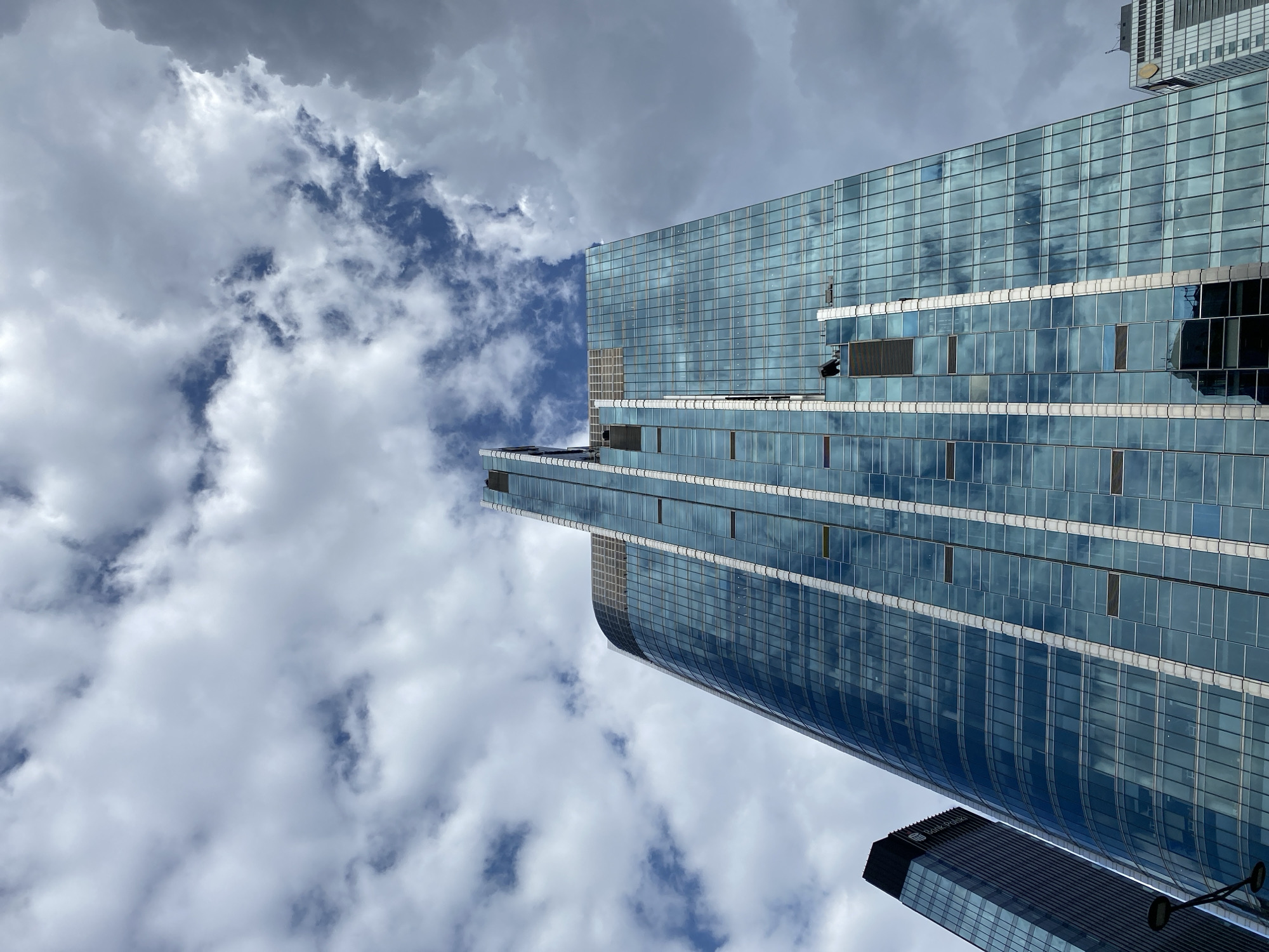 An upward view of the modern glass skyscraper SKYLIN on a cloudy day in Warsaw, Poland. The building, characterized by its blue glass facade reflecting the sky and clouds, features vertical white accents. The top of the skyscraper has a distinctive angled spire. To the left, a portion of another, darker skyscraper with a black pyramidal top is visible, along with a street lamp. To the right, the upper section of a white and gray building with a distinctive logo is peeking out from behind the main skyscraper. Two workers in dark clothing are visible on the facade of the SKYLIN skyscraper, seemingly performing maintenance or cleaning tasks, one of whom is on a suspended platform.