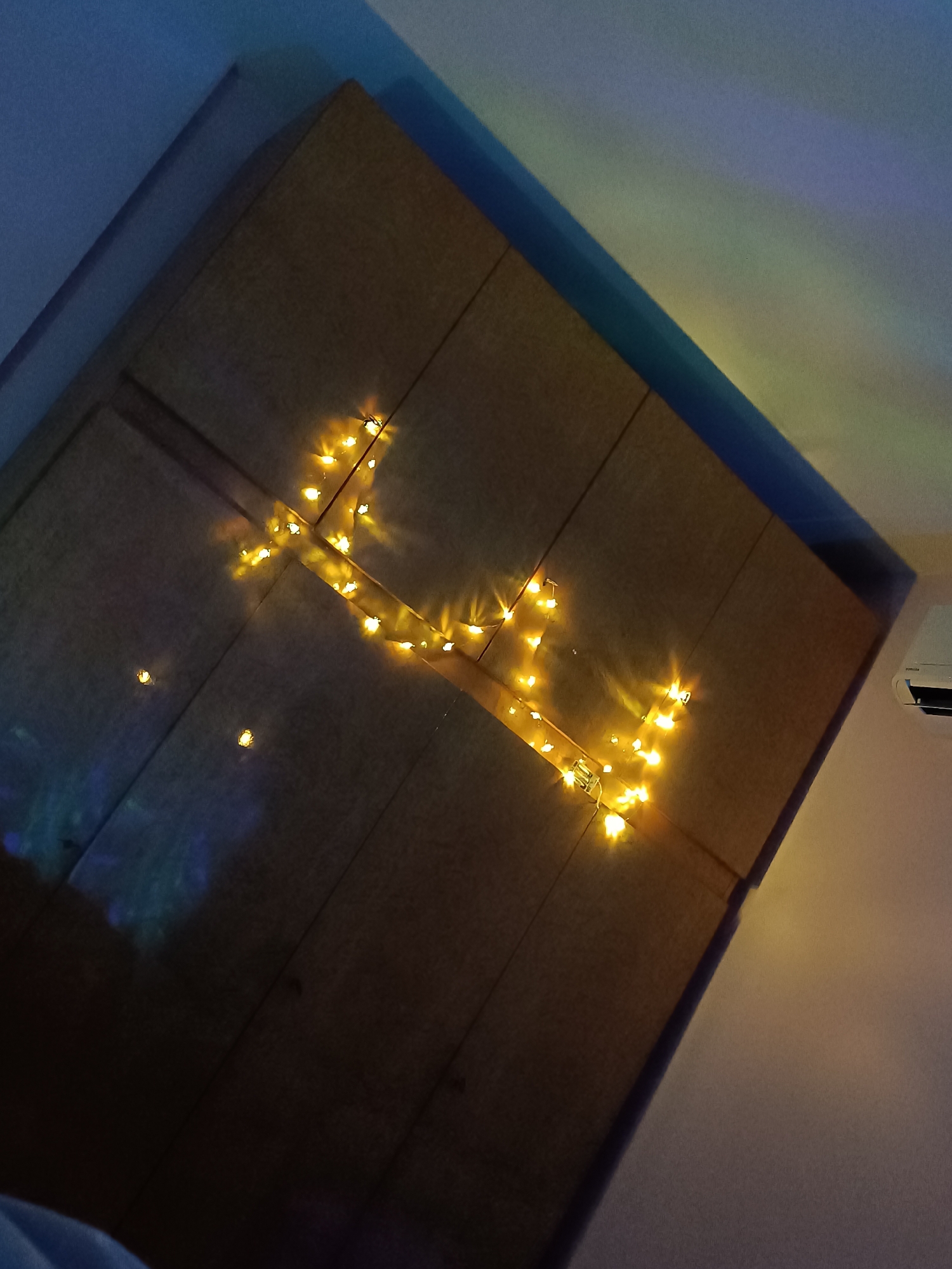 A medium shot captures a tilted view of a wardrobe or large cabinet with several vertical panels, illuminated by a string of fairy lights arranged in an irregular, non-representational pattern. The light appears warm yellow and is concentrated primarily on the right side of the object. The wardrobe is placed against the backdrop of a wall, which is light at the bottom and fades into a darker shade at the top. A corner is visible at the top-left, which shows a dark blue section of the wall. An air conditioning unit is partially visible on the right side of the image. The location is Cherkovitsa, Bulgaria.