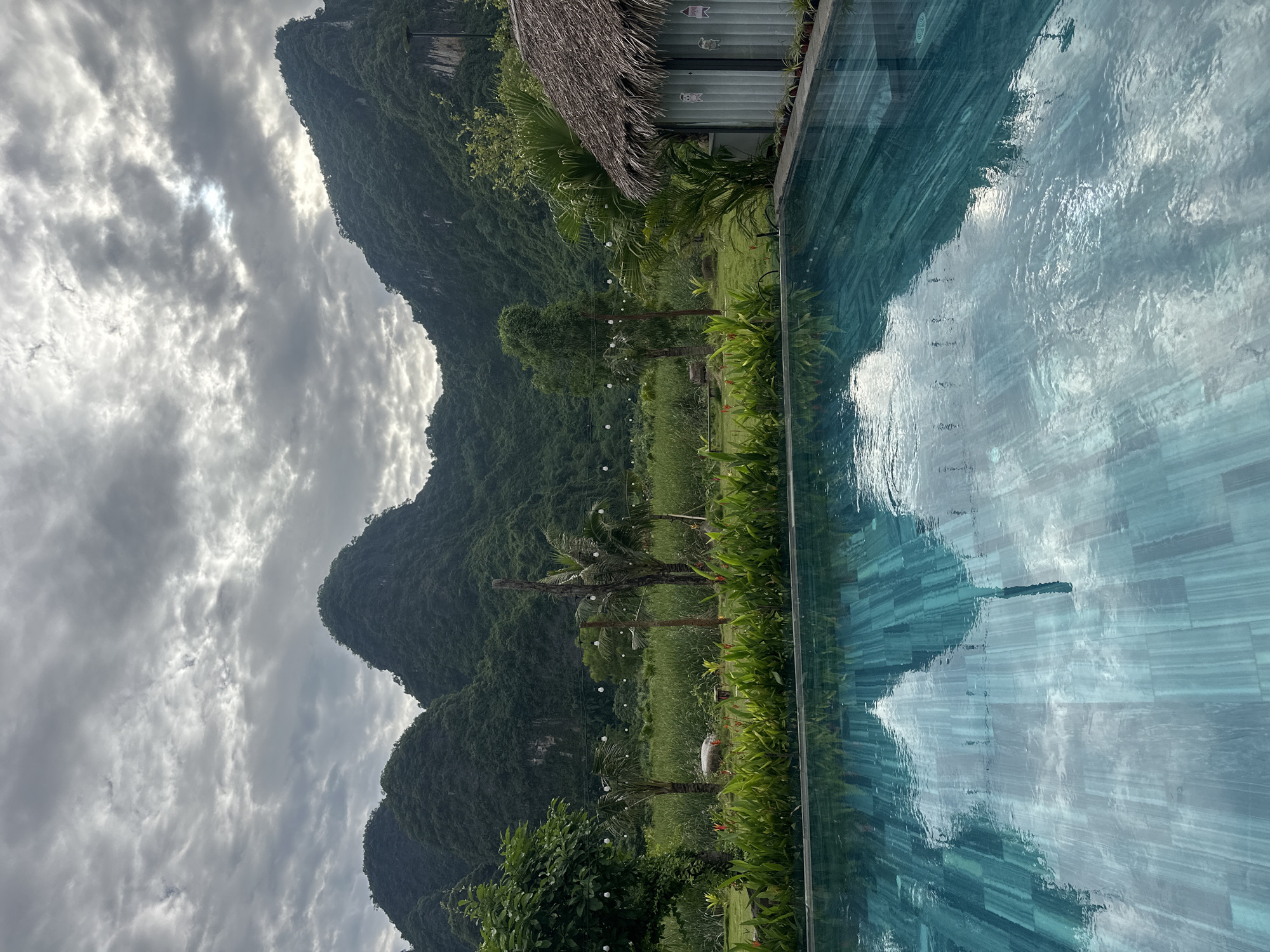The image depicts a tranquil outdoor scene with a swimming pool in the foreground, reflecting the sky and surrounding landscape. The pool features a turquoise-tiled bottom. Beyond the pool, a lush green lawn is visible, bordered by a row of vibrant green plants and red and orange flowers. In the mid-ground, a small, rustic hut with a thatched roof and corrugated metal siding is situated to the right. Tall green grasses and palm trees dot the landscape. In the background, a series of majestic, verdant mountains rise against a cloudy sky. Strings of lights are strung across the landscape, suggesting an evening setting or an area prepared for illumination. The overall atmosphere is serene and natural, likely set in a resort or rural area within Nam Hoa Lư Ward, Vietnam. No people are present in the frame.