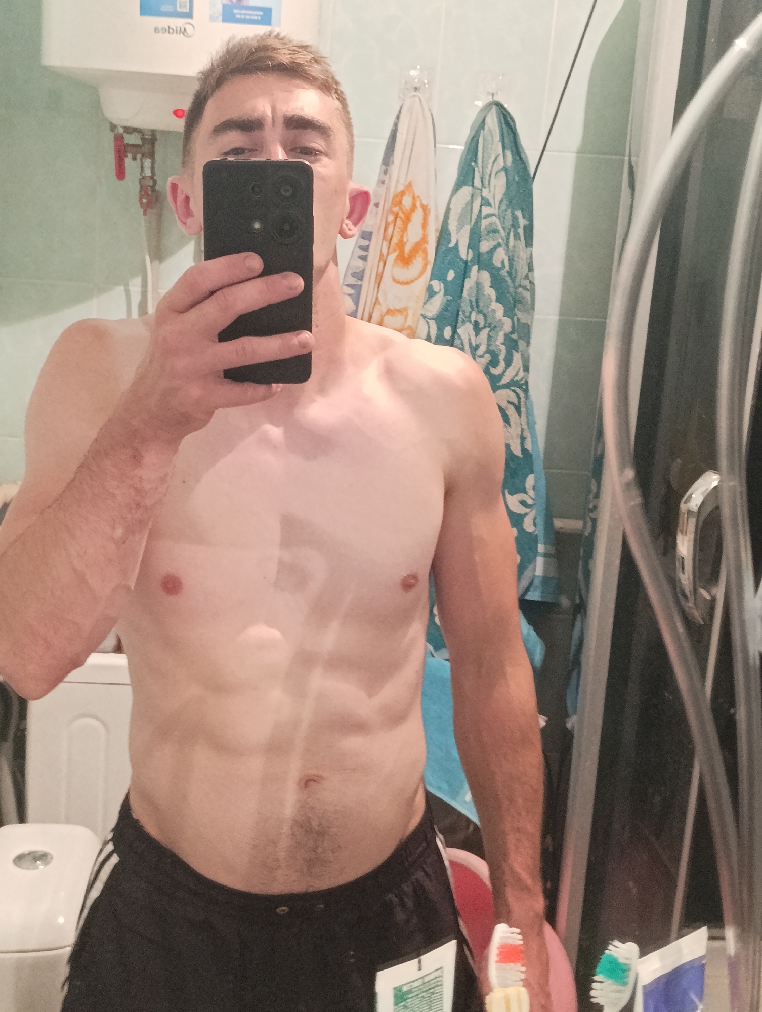 A shirtless man with short brown hair and visible abdominal muscles is taking a mirror selfie in a bathroom. He is holding a black smartphone in his right hand, obscuring his face. He is wearing black athletic shorts with white stripes. In the background, there are two towels hanging on the wall, a white tank or storage unit, and a part of a shower door. There are also toothbrushes in the foreground. The image appears to be taken in Fastiv, Ukraine.