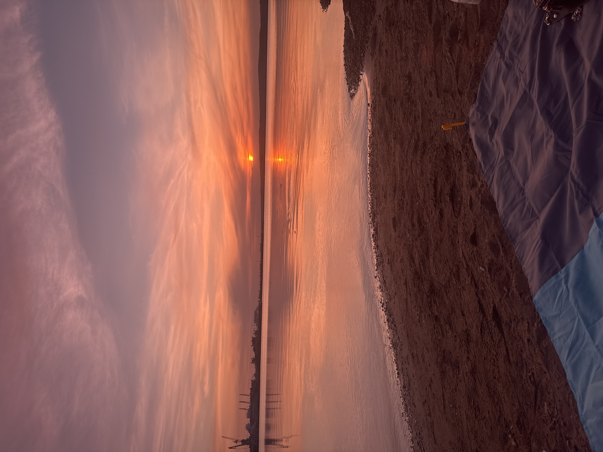 The image shows a sunset over water, likely a river or lake. The sky is filled with pink and orange clouds. The sun is visible on the horizon, reflecting off the water's surface. A shoreline runs along the right side of the image, with a beach visible. On the beach, there's a piece of cloth, possibly a blanket, and a small yellow object. In the distance, on the opposite side of the water, there are structures that may be part of a harbor or industrial area. This is likely Chaikovsky, Russia, given the industrial nature of the distant landscape along the water's edge.