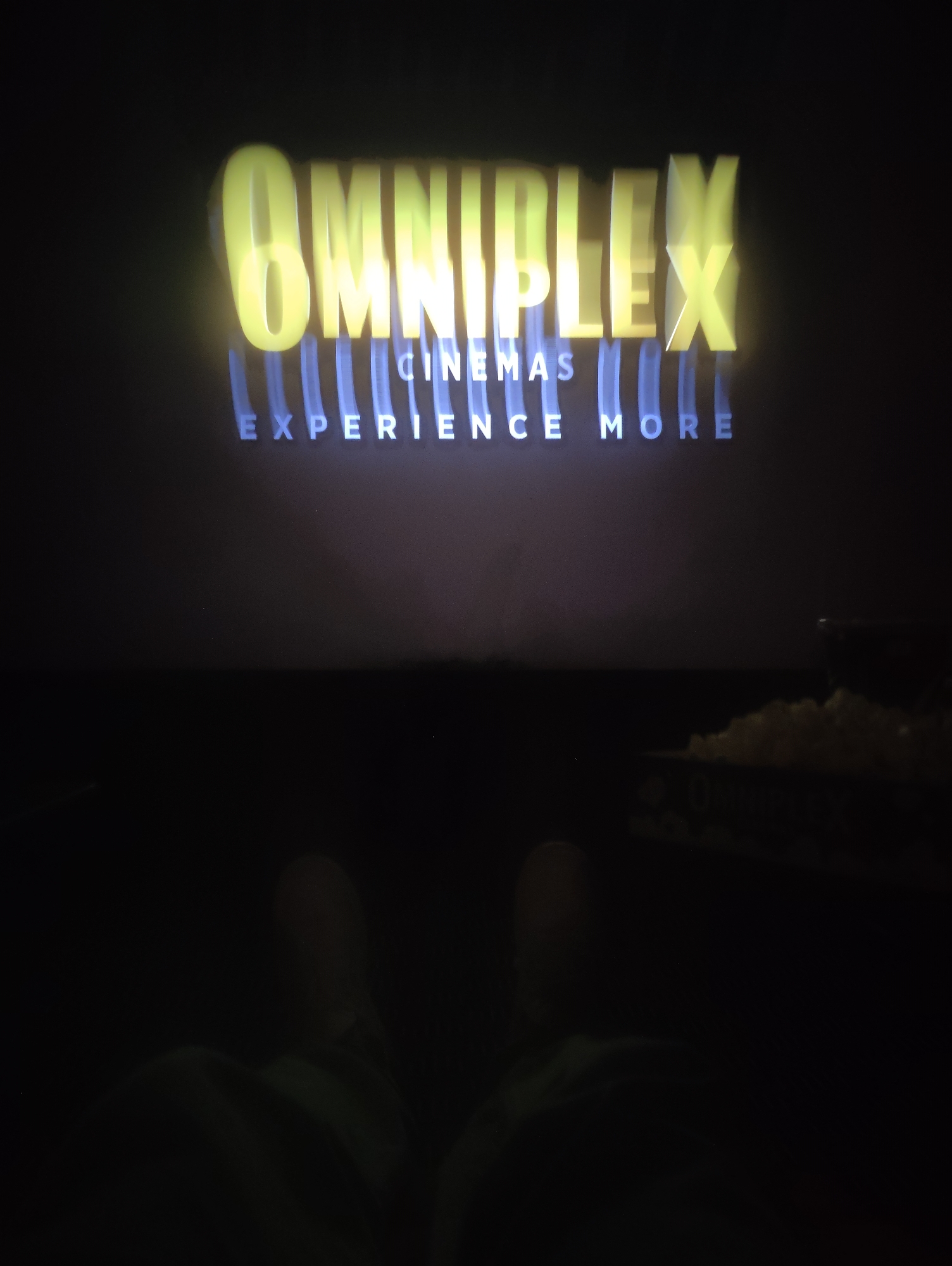 The image shows a view from inside a dark movie theater at an Omniplex Cinemas location, possibly in Ipswich, United Kingdom, as indicated by the Omniplex logo displayed on the screen. The screen displays the theater's logo: "OMNIPLEX CINEMAS EXPERIENCE MORE" in yellow and blue letters. In the foreground, there are two indistinct shapes that appear to be a person's knees or legs, and a container of popcorn can be seen to the right. The overall lighting is dim, with the main light source being the screen.
