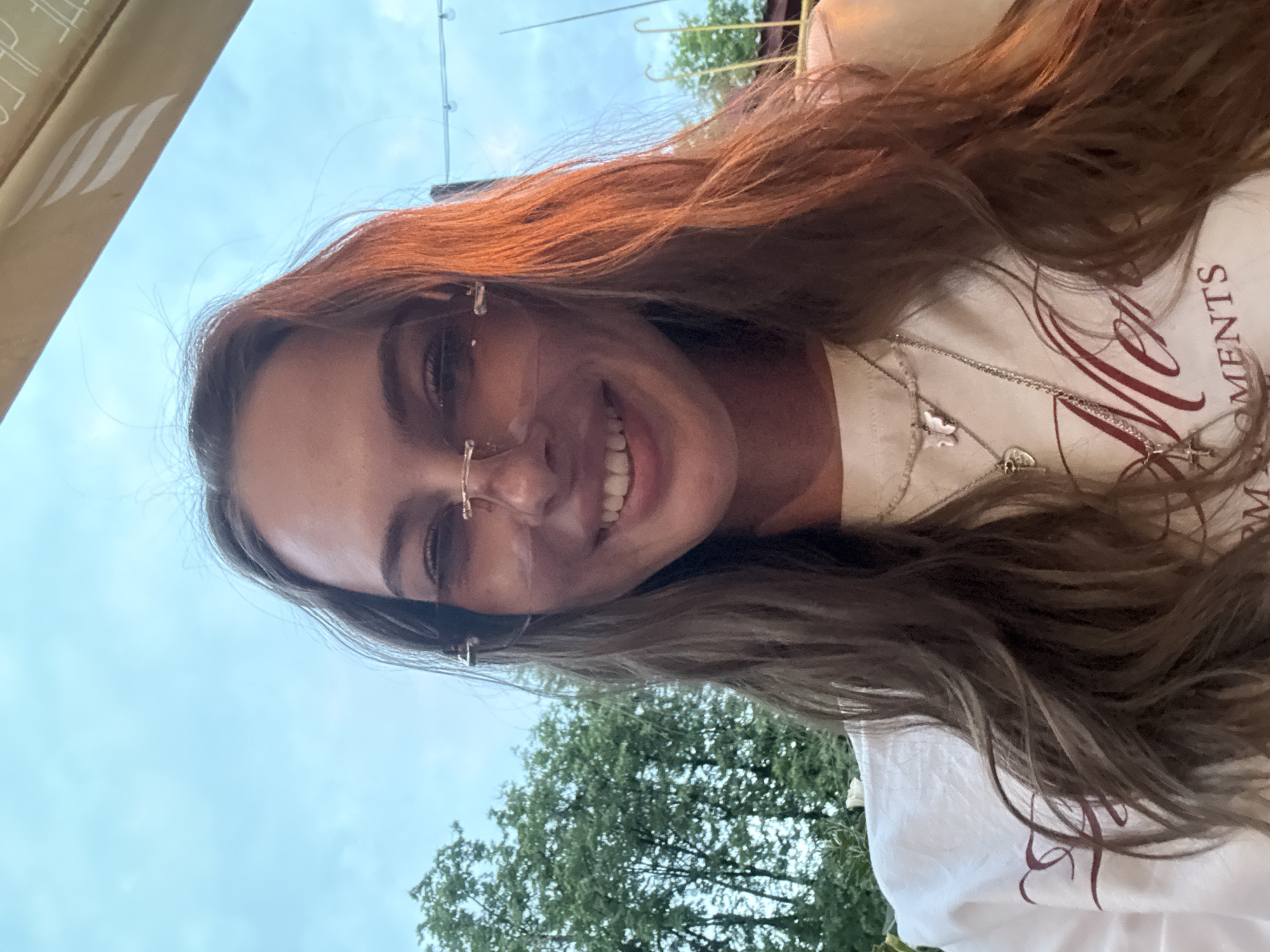 The image presents a head-and-shoulders portrait of a young adult woman, taken in a selfie style, situated in an outdoor setting in the city of Perm, Russia. The woman has long, wavy brown hair with distinct reddish-auburn highlights, particularly visible on the right side. She wears rimless sunglasses with light-tinted lenses. Her expression is a wide smile, revealing her teeth, and she is looking directly at the camera. She is dressed in a white t-shirt that features partially visible reddish-brown script lettering, spelling out "Men" and "OMENTS," likely part of the word "Moments." Around her neck, she wears multiple silver-toned necklaces: one with a pale-colored butterfly pendant, another with a heart-shaped pendant, and a third with a cross pendant. The background consists of a clear blue sky and a significant amount of green foliage from trees on the left side, indicating a natural or park-like environment. To the upper right, a section of a light-colored, possibly beige or golden, awning or canopy is visible, bearing a white, stylized emblem. Faint string lights are discernible in the distance, suggesting a patio or outdoor seating area. The lighting suggests either daytime or early evening conditions.