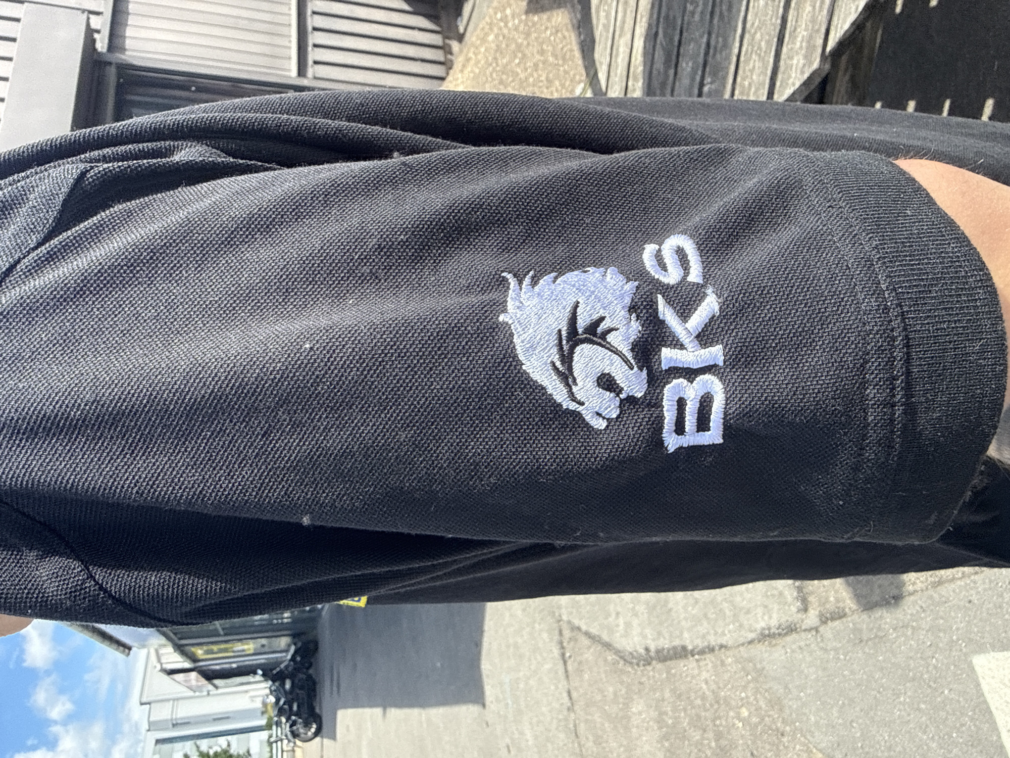 A person is shown wearing a black polo shirt with a white embroidered logo on the sleeve. The logo depicts a stylized lion's head above the letters "BKS". The background suggests an outdoor setting with a paved walkway, a building with a metal facade, and a wooden structure. The image appears to have been taken in Tewkesbury, United Kingdom, based on the context provided.