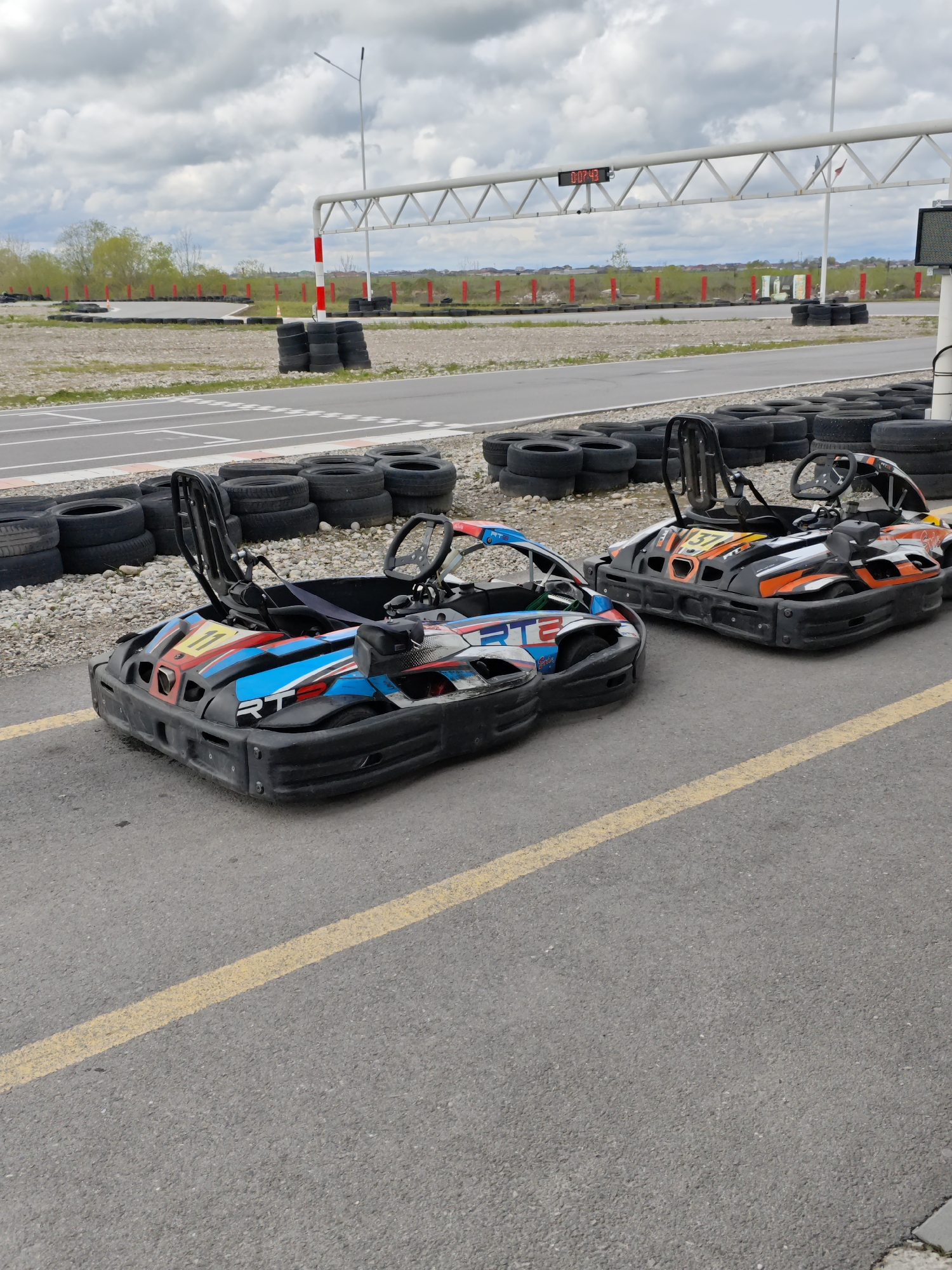 Two go-karts, one blue and one orange, are parked on a gray asphalt surface with a yellow line. Stacks of tires line the edge of the track. In the background, there is a metal archway with a digital timer. The location is an outdoor go-kart track. There are no visible people. The city is Kishpek, Russia.
