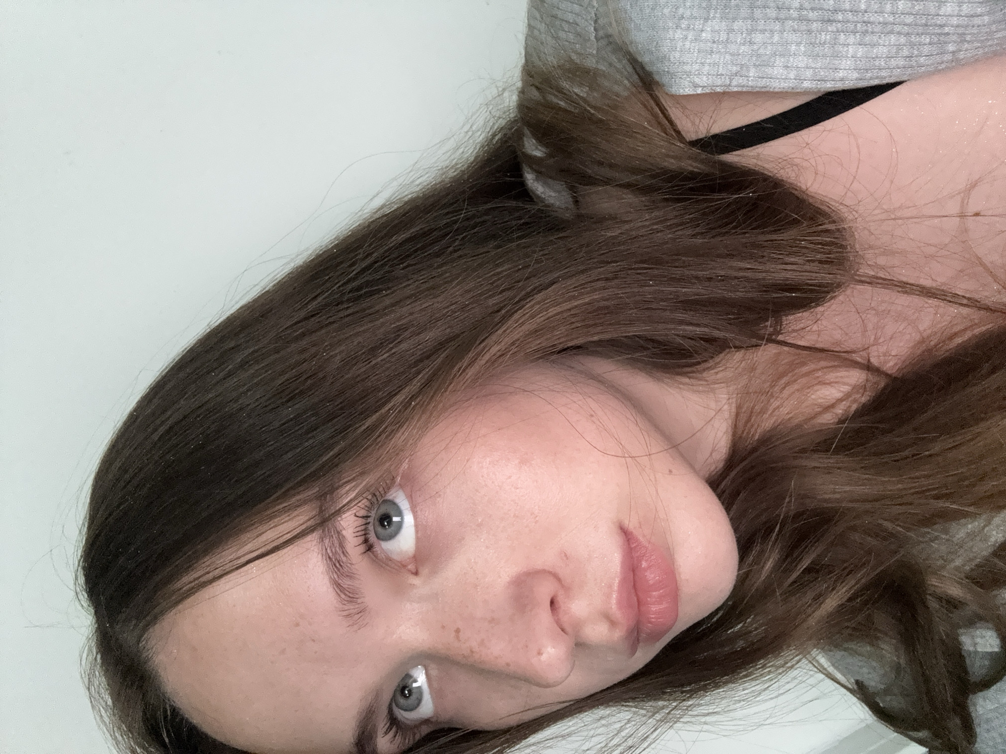 A young woman with brown hair and blue eyes looks up and to her left. She has freckles across her nose and cheeks. She is wearing a gray cardigan over a black bra. The background is a plain white wall.