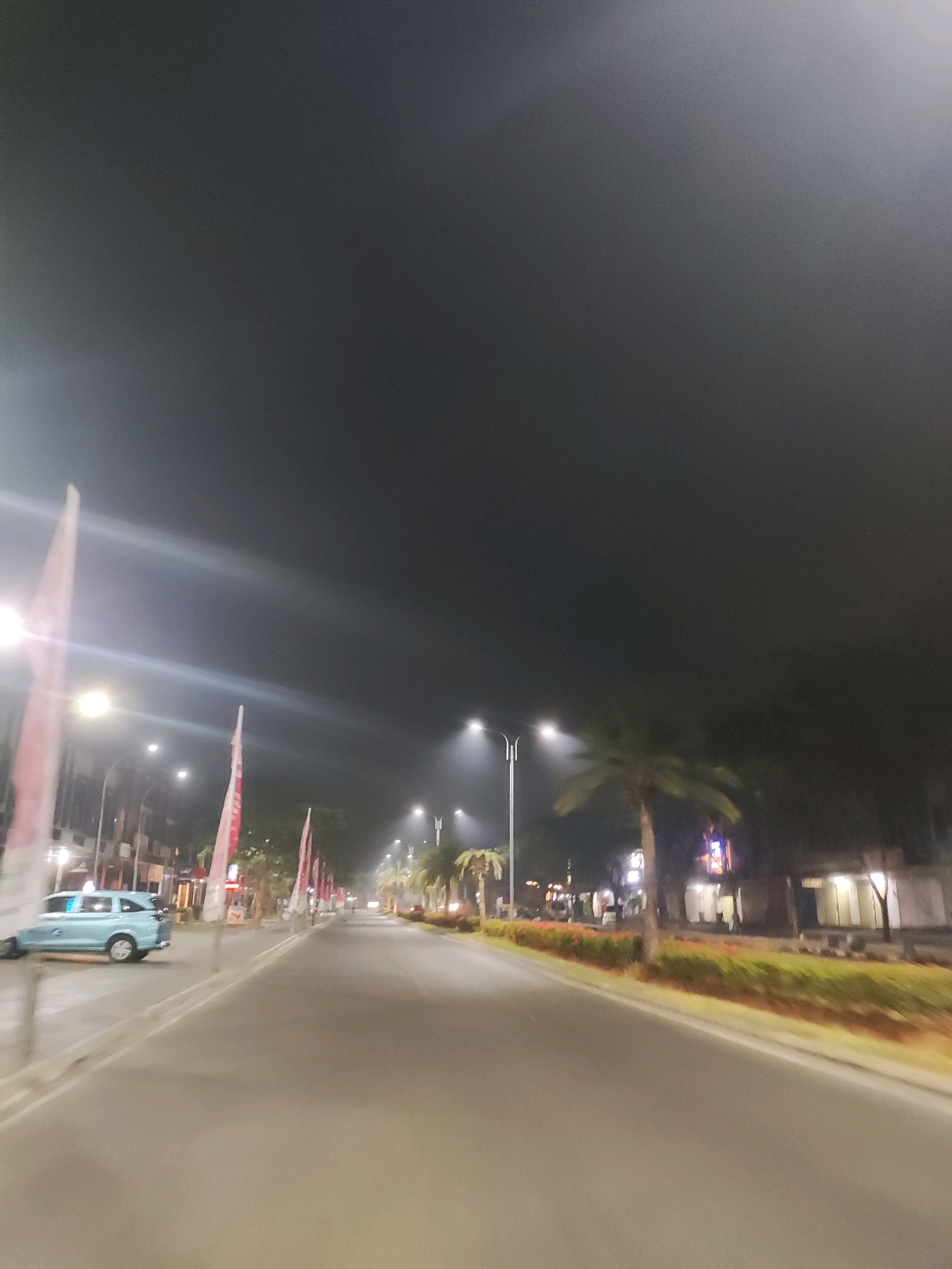 A nighttime shot captures a street scene in Telagaasih, Indonesia. The sky is a deep, hazy grey, suggesting either a cloudy night or light pollution. A street lamp on the right side of the road emits a bright glow, illuminating the misty air. To the left, a light blue vehicle is parked on the side of the road, its headlights off. Several red and white flags are prominently displayed along the sidewalk, some appearing to be Indonesian flags. Palm trees line the right side of the street, with lush green foliage bordering the road. Buildings are visible in the background, with some windows lit up, indicating that they are occupied. The road itself is paved and appears to be a main thoroughfare within the city. The motion blur in the image suggests that it was taken from a moving vehicle, likely a car or motorcycle.