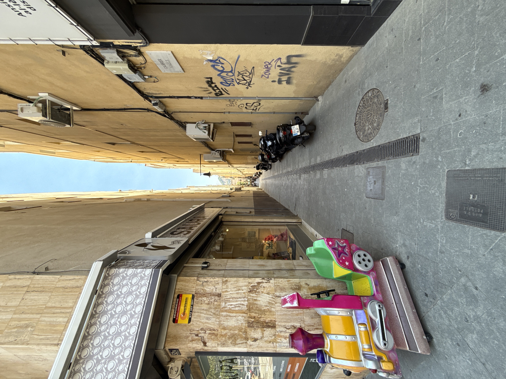 The image captures a narrow, sunlit street in Blanes, Spain. The street is flanked by tall, tan-colored buildings on both sides, with one building exhibiting a travertine facade and the other a yellow-painted facade. In the foreground, a colorful child's coin-operated toy train, painted yellow, pink, and green with decorative stars, is parked on the gray paved sidewalk.
On the left side of the street, the travertine-clad building has a shop with a large window displaying clothing. A sign on the building reads "SAURA" with a tagline below, and a yellow security sticker with "SEGURIDAD" and a phone number is visible. The shop has a patterned awning above the window.
The yellow-painted building on the right side of the street is adorned with graffiti, including the words "BIFF," "STOP," "ZOWER," and "IVAR," as well as other indecipherable tags. Various electrical conduits and a metal street sign reading "CARRER VALLS" are mounted on this wall, along with two air conditioning units and electrical boxes.
Further down the street, several black scooters are parked along the right side. The street itself is paved with gray stone slabs and features a drainage grate running along the center. A round manhole cover is visible in the pavement towards the right side of the image. The sky above is clear blue. No people are present in the immediate scene.