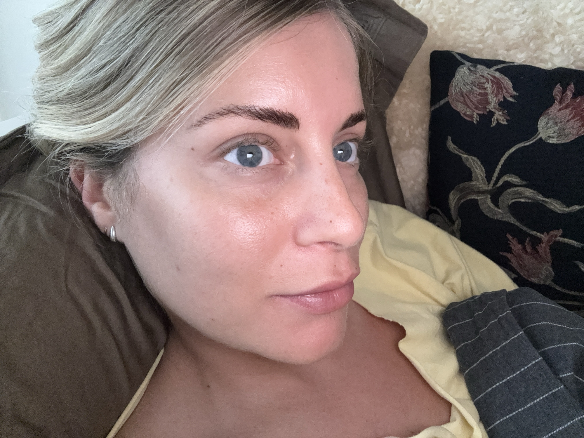 A close-up shot shows a fair-skinned woman with blonde hair pulled back from her face, resting against a brown pillow. She has blue eyes and defined eyebrows. She is wearing a pale yellow top. To her right is a black cushion with a floral pattern. A striped grey fabric also rests on the yellow top. The location cannot be determined from the image alone and Paris, France, cannot be identified.