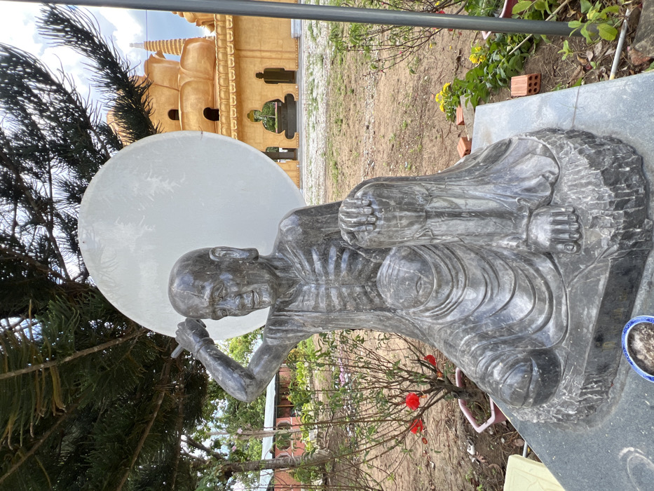 The image depicts a dark grey stone statue of an emaciated, seated figure, likely an ascetic or Buddhist monk, located outdoors within a religious complex in Đăk Mar, Vietnam. The statue, positioned centrally, shows a male figure with visible ribs, a serene expression, and a slight smile. The figure's right arm is raised, holding an elongated object (possibly a staff or brush) over its head, while the left hand rests on the raised left knee. The statue rests on a dark grey stone pedestal.
Behind the statue's head is a large, circular, white disc, possibly a halo or a decorative element. To the right of the statue, a prominent, multi-tiered golden-yellow pagoda or temple building with distinctive lotus-petal shaped structures and spires is visible in the midground. Archways at the base of the temple reveal dark silhouetted figures or architectural details within.
Overhead and to the left, dark green foliage from a large evergreen tree partially shades the scene. The foreground and midground consist of bare earth with sparse green vegetation, including small bushes with red flowers (like a rose) to the left and yellow flowers to the right. Red bricks are scattered on the ground near the base of the statue on the right. In the immediate foreground, a small blue and white patterned bowl containing a dark substance, possibly ash, rests on the statue's base. The background features a partly cloudy sky. To the far left, a simple building with a red roof is partially visible amidst trees.