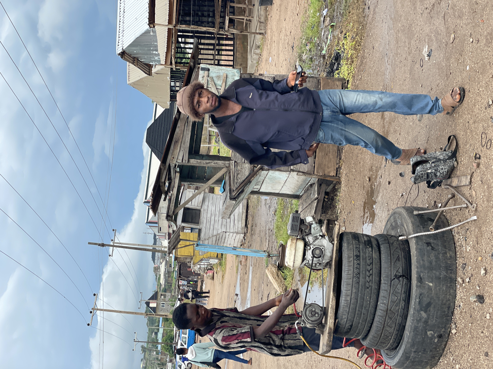 In this outdoor scene in Lokoja, Nigeria, two young men are engaged in a tire repair activity. One man, wearing a dark blue jacket, blue jeans, and a brown furry hat, stands near a small engine and a stack of tires. He holds a black smartphone in his right hand and appears to be looking towards the camera. Beside him, a patterned backpack rests on the ground. The other man, dressed in a striped shirt and dark pants, is seated and actively working on one of the tires. He is connected to a device, possibly a tire inflator, which is part of the setup that includes an engine and a metal frame holding the tires.
The setting is an unpaved, somewhat muddy area with makeshift wooden structures and corrugated metal roofs suggesting a commercial or workshop space. In the background, there are more buildings, power lines, and a general sense of a busy, somewhat rustic environment. The sky is partly cloudy, indicating it could be daytime. The overall atmosphere appears to be one of everyday work and service provision. There is no visible text in the image.