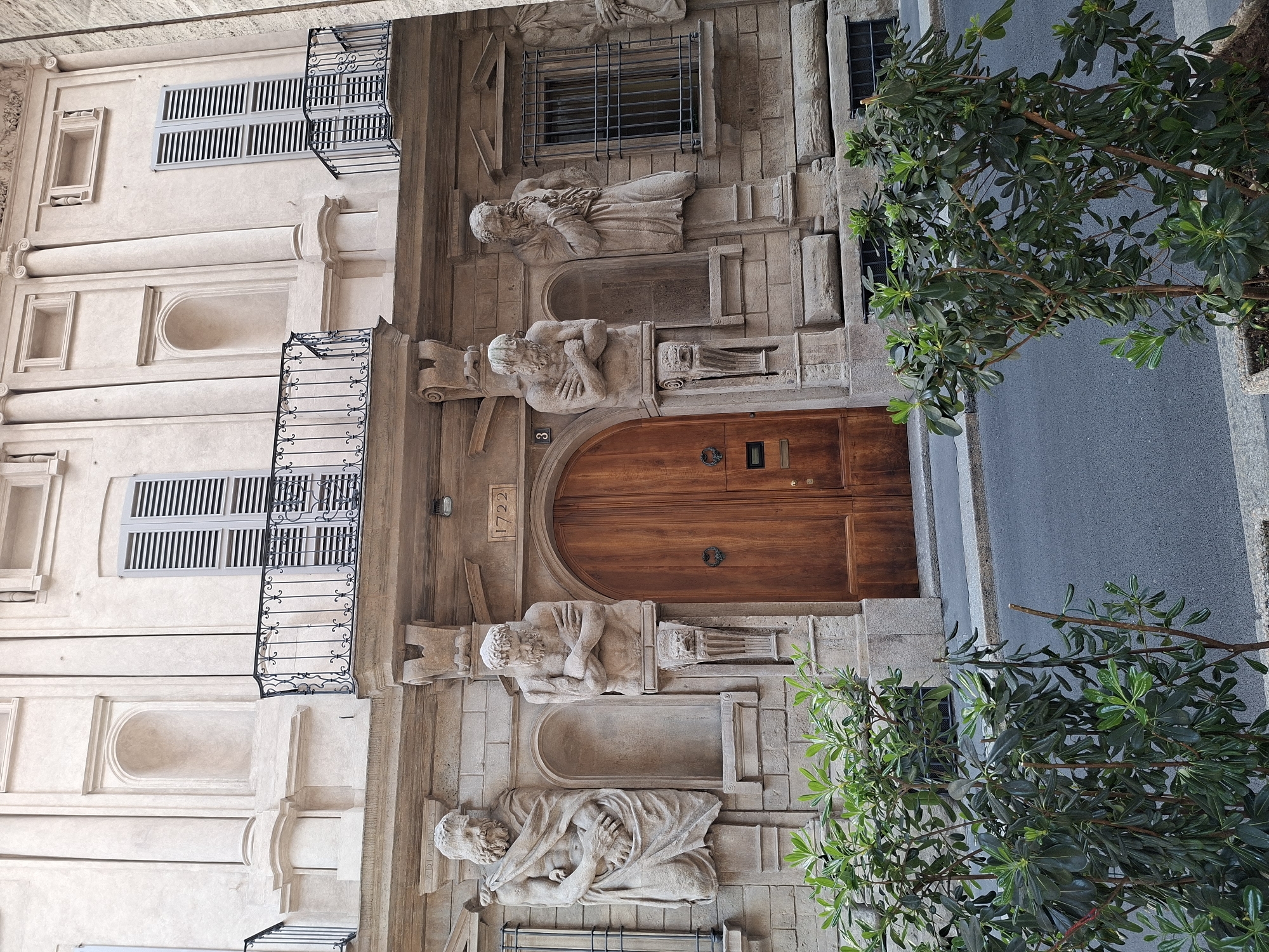 The image captures a building facade in Milan, Italy, with a prominent wooden door flanked by stone sculptures of reclining figures. The facade is detailed with ornate stonework, including a barred window above the door and decorative elements. Balconies with black iron railings are visible on the left side of the building. The number "8" and the year "1722" are inscribed near the door. A portion of a tree with green foliage is visible on the right side of the frame, partially obscuring the sidewalk or street. The building's architecture suggests a historic or classical style.