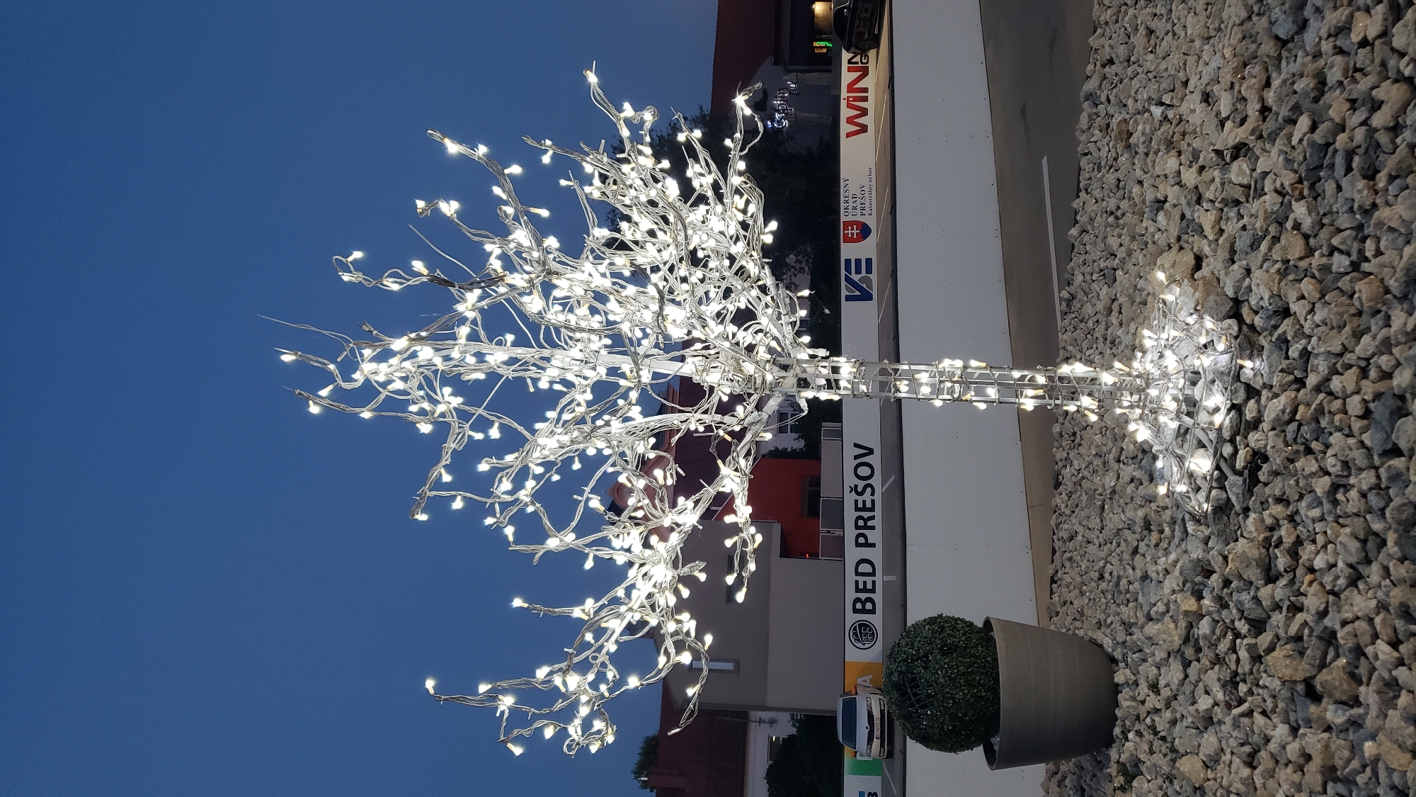 A decorative, artificial tree illuminated by numerous white LED lights stands in a gravel-covered area at dusk. The tree has a wireframe structure wrapped in lights, forming a trunk and many branching limbs adorned with small white bulbs. To the left of the tree, a dark gray pot contains a spherical, topiary-style bush. In the background, a white barrier wall is visible, displaying several logos and text. One prominent logo reads "BED PREŠOV". Behind the wall, buildings with red and gray facades are dimly lit. The sky above is a deep blue, characteristic of twilight. The scene appears to be an outdoor public space, possibly a plaza or streetscape in Prešov, Slovakia.