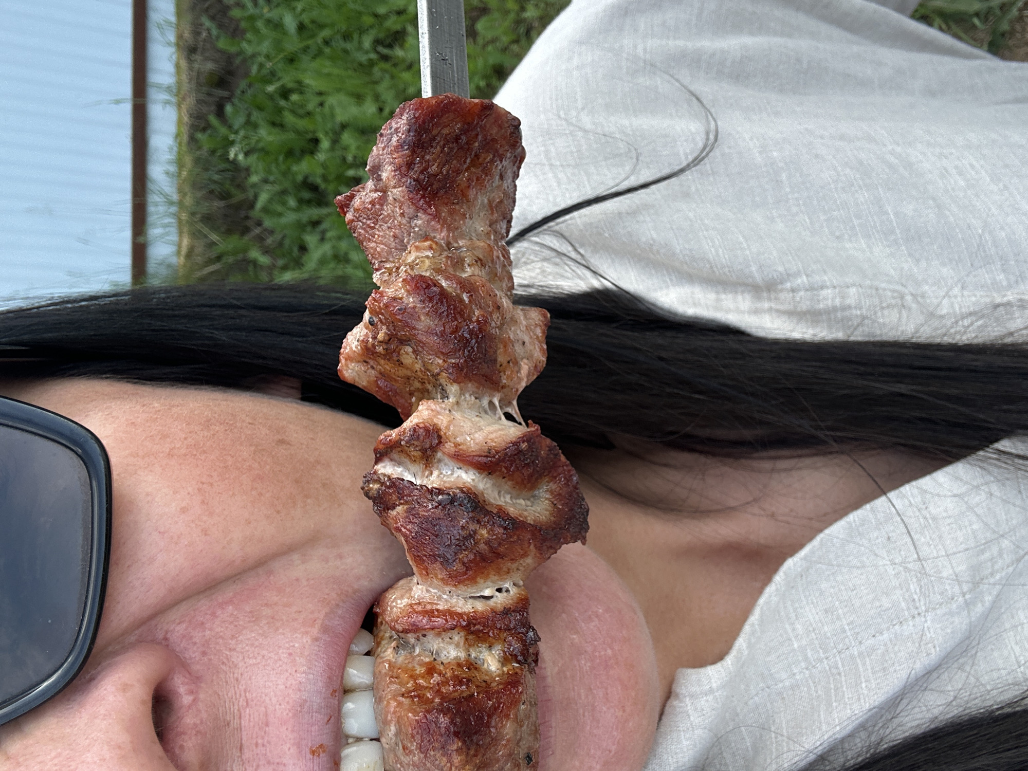 A close-up shot shows a person's face from the nose down, wearing dark sunglasses and a light-colored, textured shirt. The person is biting into a piece of grilled meat on a metal skewer. The meat is browned and appears juicy with visible layers of fat. The background is blurred, showing green grass and a light-colored structure that may be a building or fence. The setting appears to be outdoors.