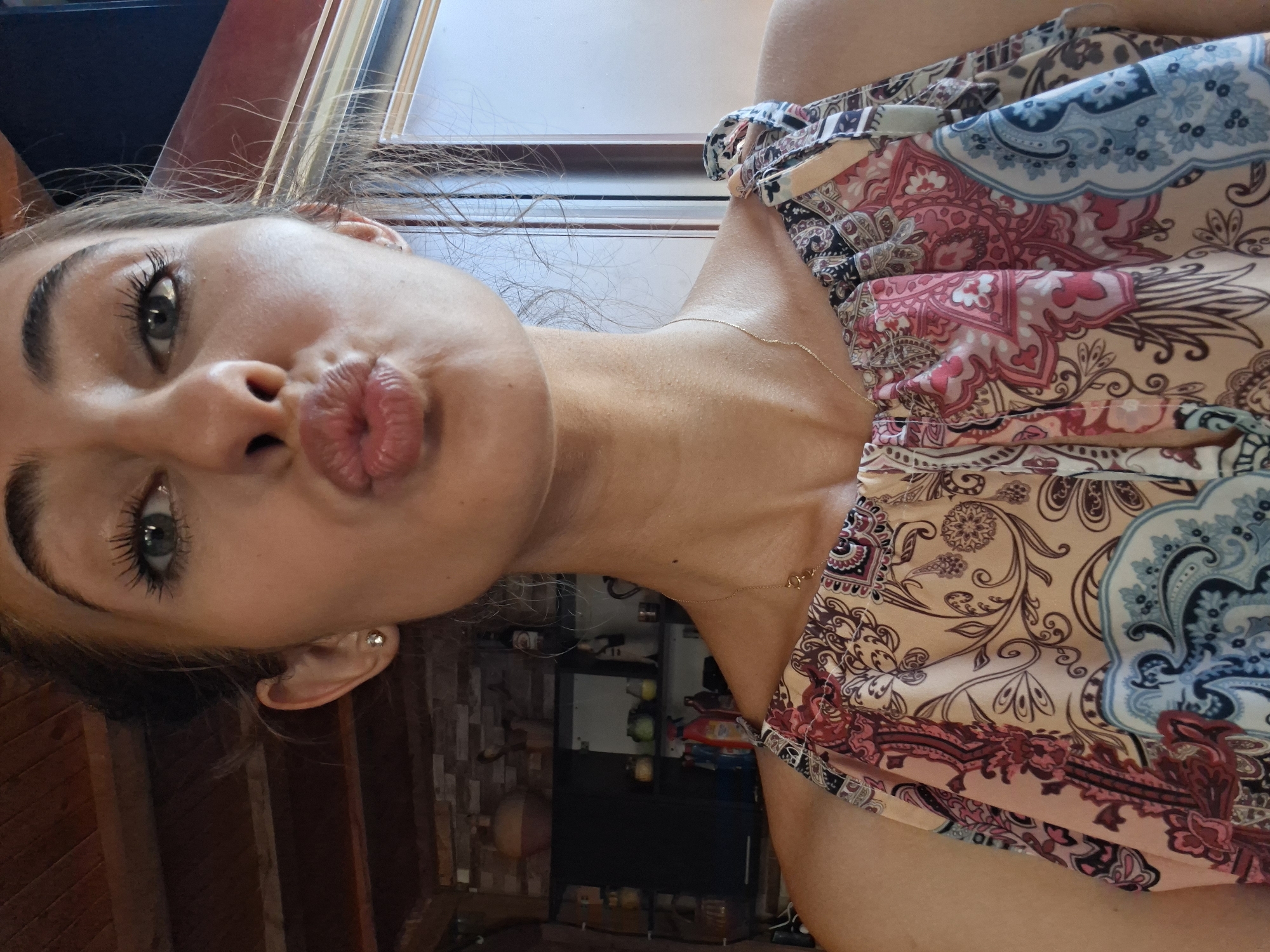 A close-up, forward-facing selfie of a young woman with light skin, brown hair pulled back, and green eyes. She is puckering her lips and wearing a tan, pink, and blue patterned sleeveless dress. A thin gold necklace is visible around her neck. The background shows a wooden ceiling and a blurred interior with shelves containing various objects. The lighting suggests an indoor setting, possibly during the day.