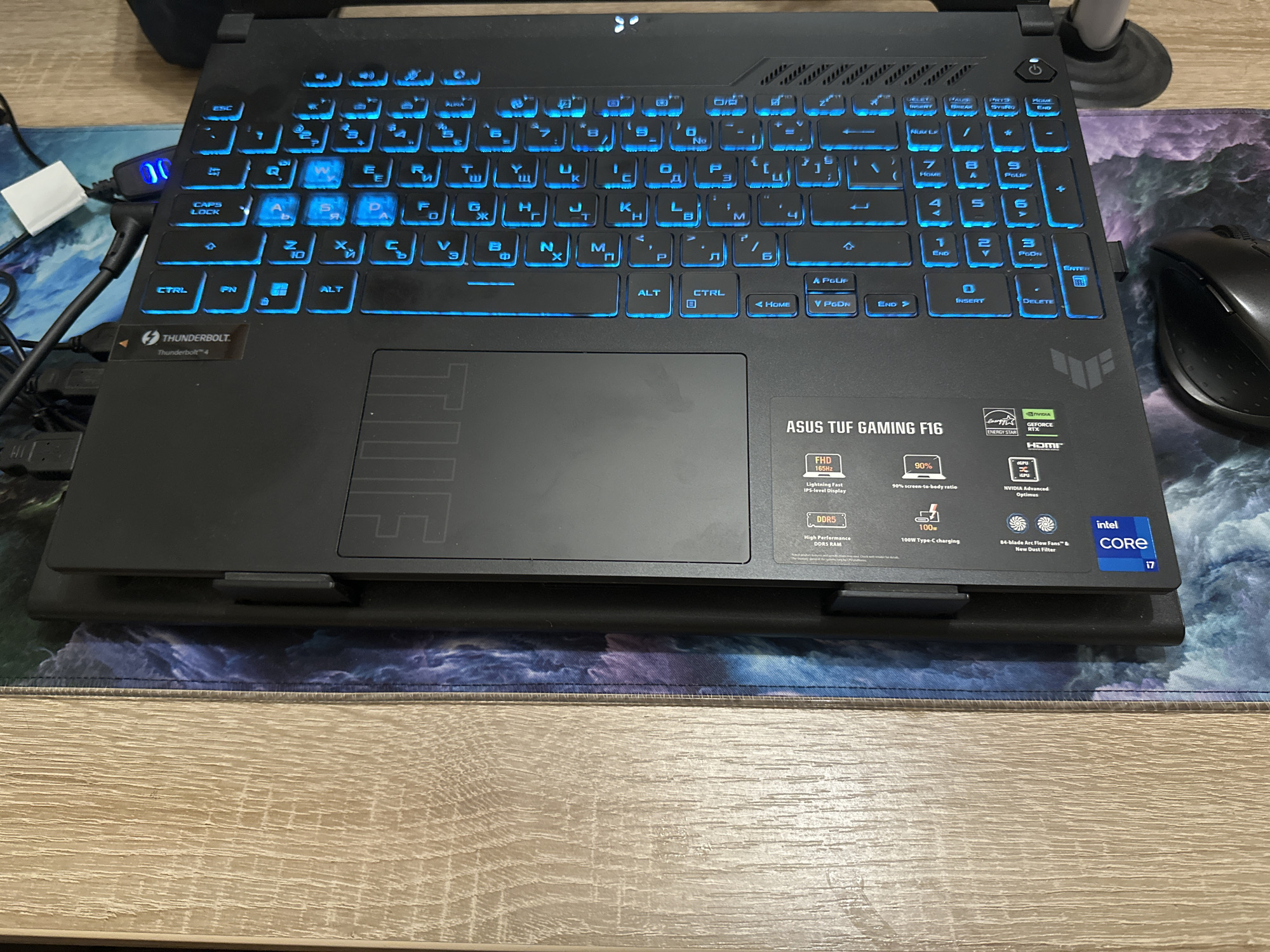 The image displays a top-down, slightly angled view of a black ASUS TUF GAMING F16 laptop oriented vertically. The laptop's screen side is towards the left edge of the frame, and the keyboard and trackpad are towards the right.
The laptop features a black chassis with a bright blue backlit keyboard, displaying both English (QWERTY) and Cyrillic character layouts on the keys. A large, dark grey trackpad is positioned centrally below the keyboard. On the palm rest, several stickers are visible, including "Thunderbolt 4" near the upper-left of the keyboard, an "intel core i7" logo, an "NVIDIA GeForce RTX" logo, and various feature highlights such as "DDR5" RAM, "FHD 165Hz" display, "100W Type-C charging," "90% screen-to-body ratio," and "64-blade Arc Flow Fans" with an "Anti-Dust Filter." The "ASUS TUF GAMING F16" model name is also prominently displayed on a label on the right palm rest.
Along the top edge of the laptop (which is the left side of the image frame), several black USB cables are connected to ports. One cable leads to a small, dark blue USB hub or adapter that emits a distinct blue light, indicating power or activity. A small white tab or cable is also visible near this hub.
To the lower right of the laptop, a black computer mouse, appearing to be an ergonomic right-handed model, is partially visible.
The laptop and mouse rest on a large gaming mouse pad with a dark blue and purple abstract design, resembling swirling water or clouds. The mouse pad has a black stitched edge. To the left of the mouse pad, a light brown wooden surface, likely a desk, is visible, displaying a prominent vertical wood grain pattern. The scene is well-lit, suggesting ambient overhead lighting, with no significant shadows present. No individuals are visible in the frame. The setting is an indoor environment in Unknown, Unknown.