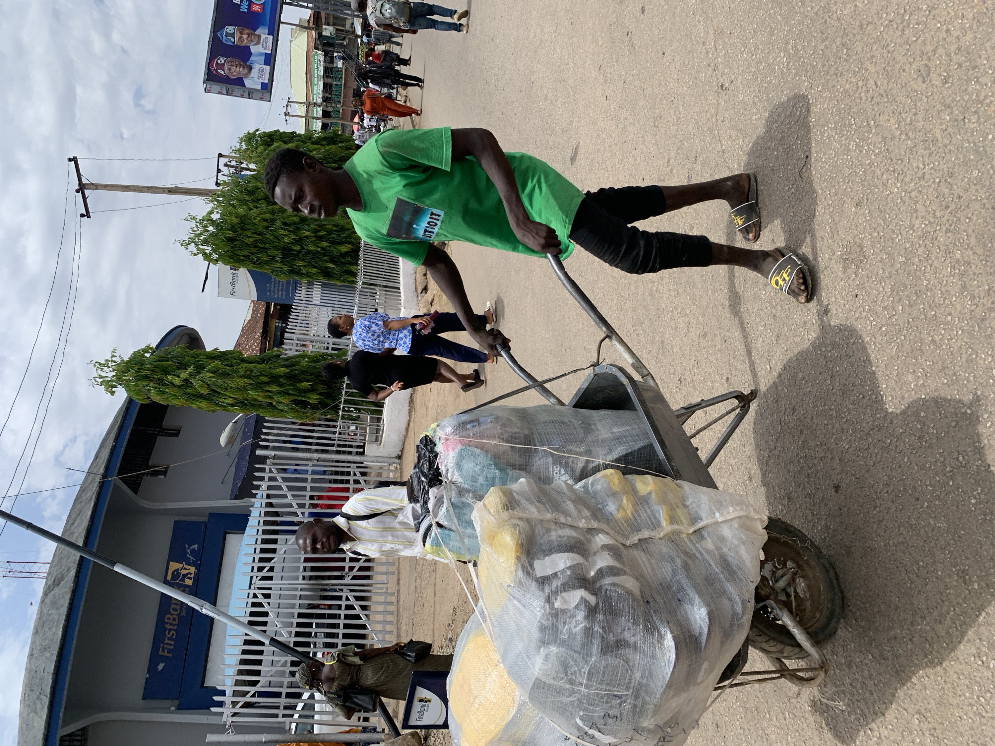 The image captures a man wearing a green shirt and black pants pushing a wheelbarrow filled with wrapped items on a paved street. He is wearing "Off-White" branded sandals. In the background, other people are walking, and there's a building with a "FirstBank" sign visible. Overhead power lines are also seen. A political billboard is located on a pole. The location is identified as Jos, Nigeria.