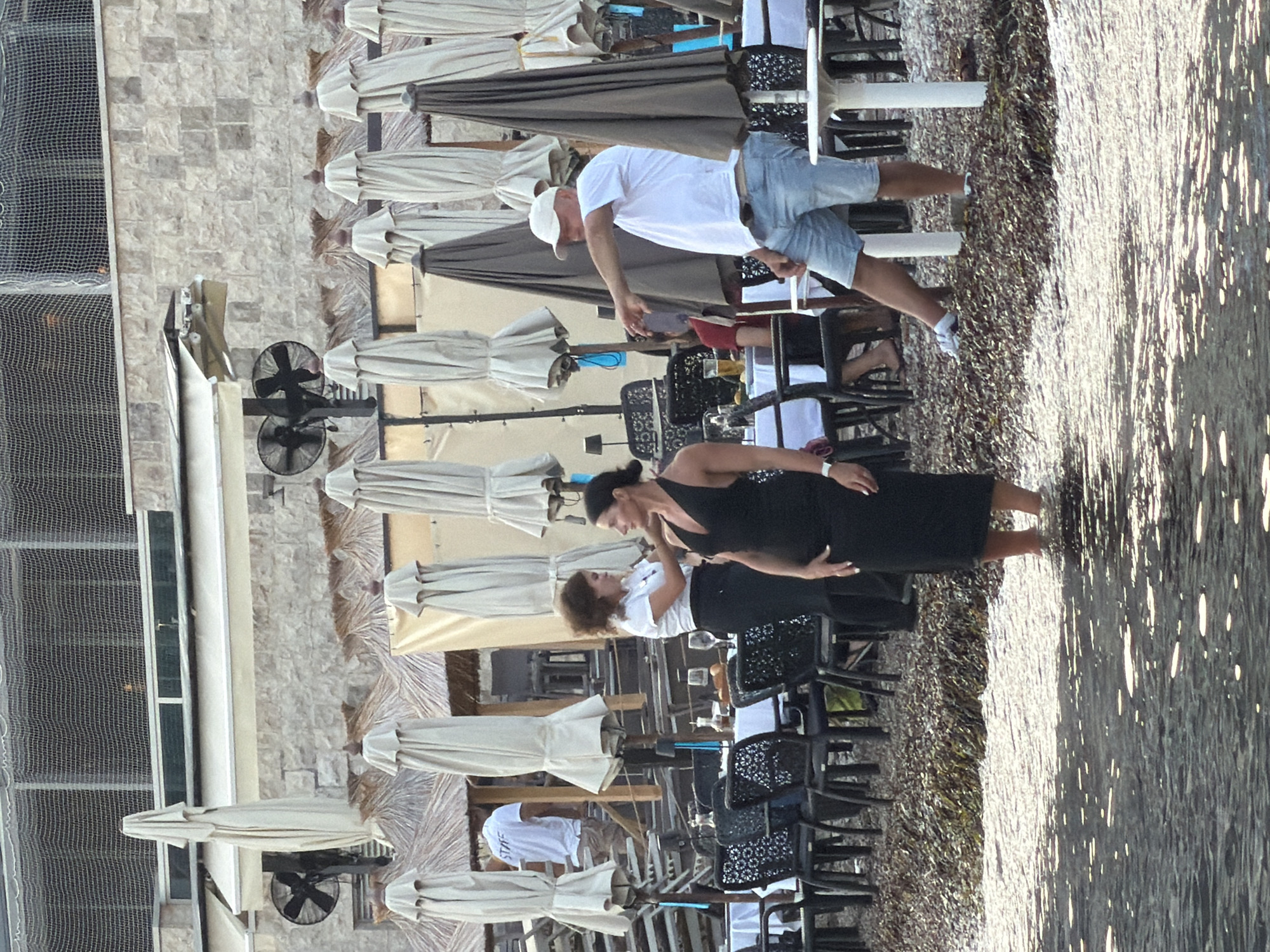 The image depicts an outdoor seating area of a restaurant or cafe, likely situated on a beach or waterfront in Budva, Montenegro. In the foreground, shallow water with reflective sunlight covers the lower part of the frame. The midground features a pebbly ground with scattered seaweed, leading to a row of tables set with white tablecloths and chairs. Several closed and open umbrellas are visible, providing shade.
In the midground, three individuals are the primary subjects. On the left, a woman wearing a black dress stands with her hands on her hips, facing right. Next to her, also facing right, is a woman in a white shirt and black skirt, appearing to be a server, possibly interacting with the woman in the black dress. On the right, a man in a white t-shirt and denim shorts, wearing a white baseball cap, stands and points his left arm outwards towards the right, as if directing someone or indicating something. He is partially obscured by a large, dark umbrella.
In the background, the facade of a building constructed with stone blocks is visible, with various awnings and structures. Two large industrial fans are mounted on poles, suggesting a need for cooling in the outdoor environment. A net structure is visible overhead. A person wearing a white shirt with "STAFF" printed on the back is seen in the far background, near some stacked chairs. The overall scene conveys a relaxed yet functional atmosphere of a coastal establishment.