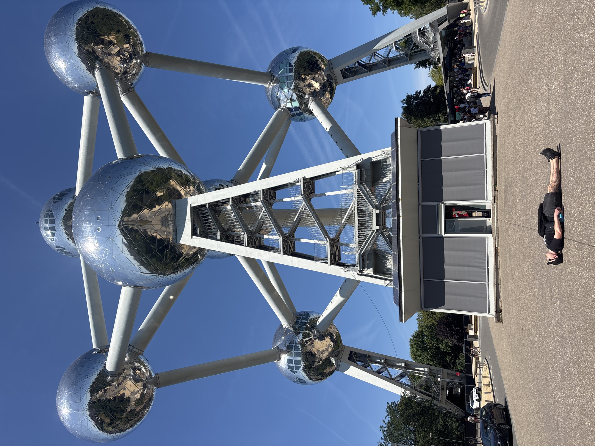 The media file displays the Atomium in Brussels, Belgium, under a clear blue sky. The iconic structure, designed to resemble a unit cell of an iron crystal, is the central subject. Its large, spherical compartments are clad in polished metal, reflecting the surrounding environment, including trees and the blue sky. Several of these spheres are interconnected by tubes, forming a prominent architectural landmark.
In the foreground, a person is lying on the asphalt surface, oriented in the same direction as the overall image which appears to be rotated 90 degrees counter-clockwise. This person is wearing dark clothing and a baseball cap. The ground is paved with asphalt and there are some vehicles visible further down the path, including a black car and a white car.
In the background, a crowd of people is gathered near the base of the Atomium, possibly waiting to enter or observe the structure. Trees and foliage are visible, indicating an outdoor setting. The presence of the Atomium, a well-known tourist attraction, suggests this is a public space, likely during daytime given the bright sunlight and clear sky. The overall atmosphere appears to be relaxed and leisurely, consistent with a visit to a famous monument on a pleasant day. No visible text is present in the image.