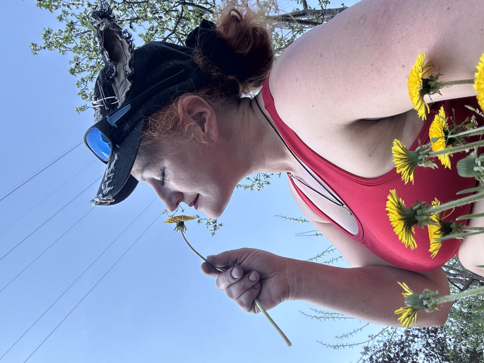 A low-angle shot depicts a fair-skinned person smelling a dandelion in Chaikovsky, Russia. The person, wearing a red swimsuit and a black cap adorned with goggles and decorative pins, holds the dandelion in their right hand. Their red hair is styled in a bun. Several dandelions bloom in the foreground. The sky is blue. Electrical wires stretch across the upper left. The scene is outdoors, during daylight, with the person positioned against the sky and a tree with sparse foliage.