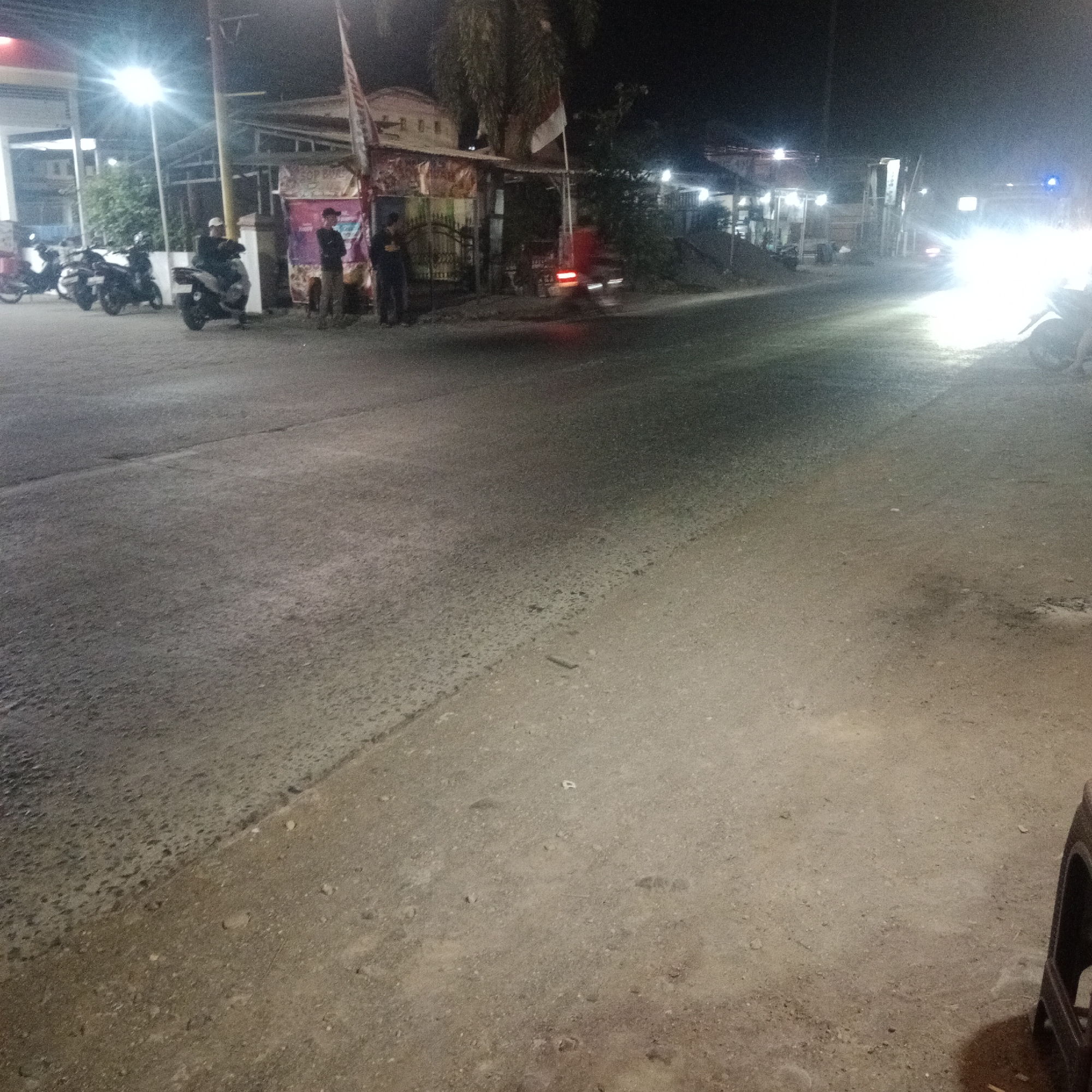 The image depicts a street scene at night in Tegowanu Kulon, Indonesia, near a gas station. Several motorcycles are parked on the left side of the road, and a few individuals are standing near a small building or kiosk. The road surface is asphalt, with some areas appearing to be gravel or dirt. Bright lights from the gas station and oncoming vehicles illuminate the scene, creating significant glare and shadows. The overall atmosphere suggests a typical evening in a semi-urban or rural area.
