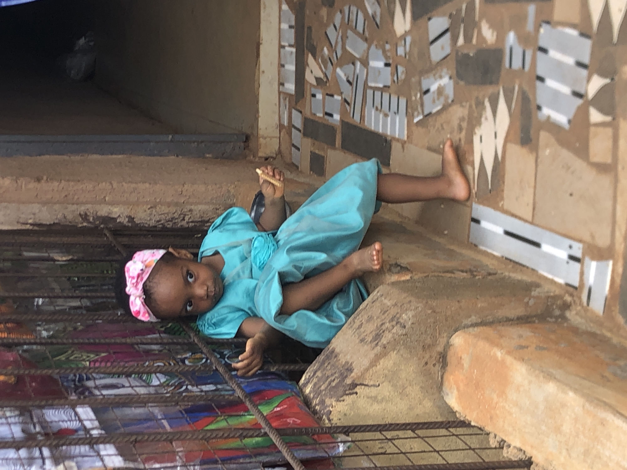 A young child, likely a girl, is pictured lying on her side, gazing directly at the viewer. She wears a light blue dress with a ruffled detail on the shoulder and a pink and white patterned head wrap. Her body is positioned horizontally across a rugged, earthen or concrete surface, which appears to be a ledge or step. In her right hand, she holds a thin, light-colored object, possibly a snack.
The immediate foreground features a dark metal grate or railing running horizontally, suggesting a barrier or window. Behind the child, a wall is decorated with an abstract mosaic of irregularly shaped light grey and white pieces, some featuring black horizontal stripes, set against a brown, textured backdrop. To the child's left, a dark, shadowed recess is visible in the background, perhaps an interior corner or opening. The natural lighting suggests it is daytime. The child's expression is one of calm curiosity. This scene is located in Ukwu-Abwa, Nigeria. No visible text is present in the image.