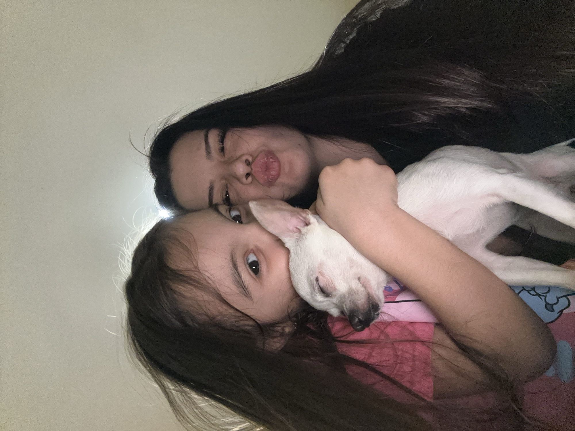 Two females and a dog are present in the image. The adult female has long, dark hair and is making a kissing face. The child has long, dark hair and is looking at the camera. The dog is white and is being held by the child. They are inside a room. The location is not identifiable as Chubra, Bulgaria.