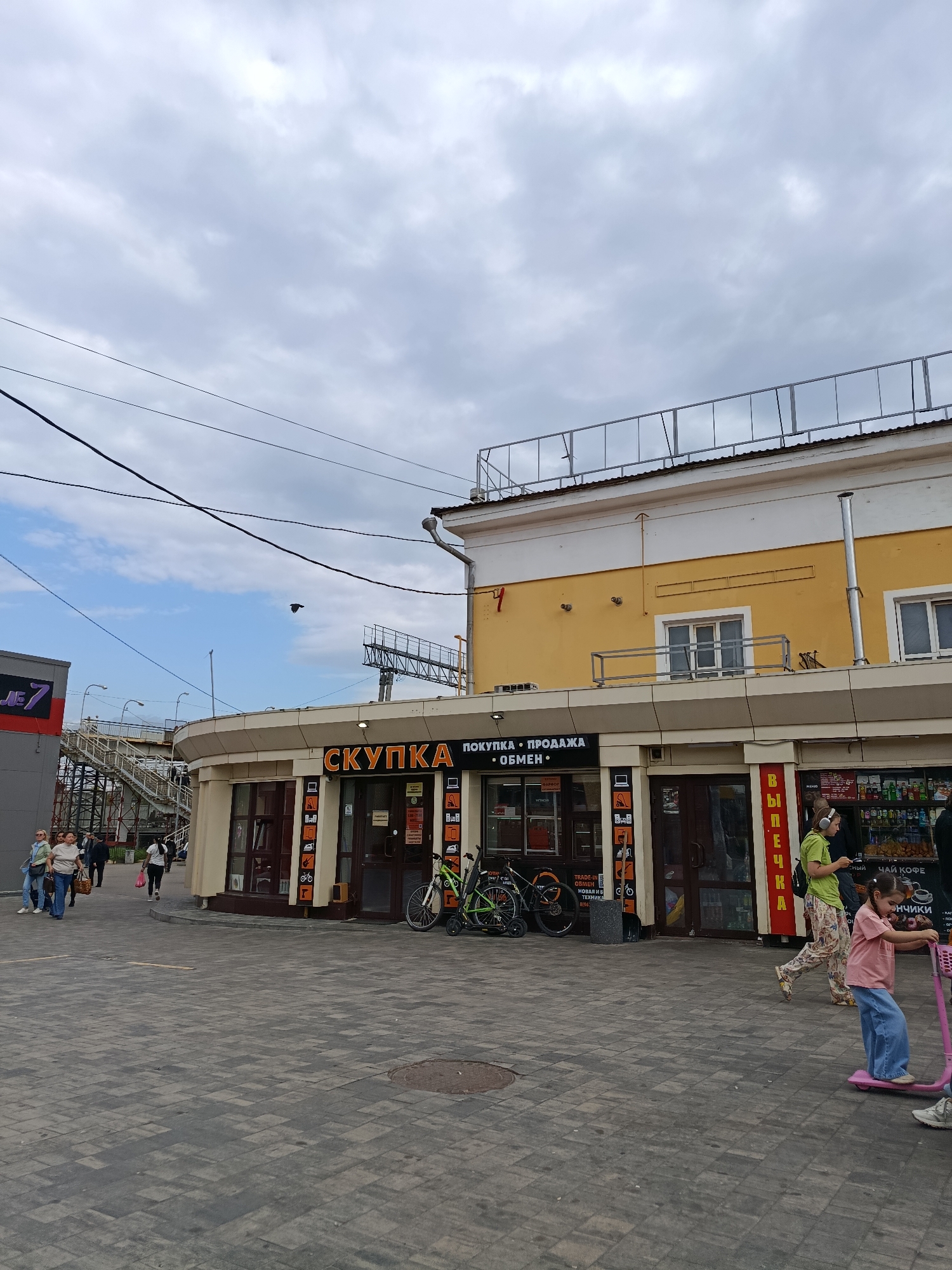 In Orekhovo-Zuyevo, Russia, a street-level view captures a curved building with a yellow upper facade and a lighter-colored, circular storefront below. The storefront features multiple glass-paneled doors and windows, with prominent signage that reads "СКУПКА" (purchase) in large orange letters, followed by smaller text indicating "ПОКУПКА • ПРОДАЖА • ОБМЕН" (purchase, sale, exchange). Several people are visible. On the left, a group of women walk along the paved area in front of the building. To the right, a woman in a green shirt and patterned pants walks past, her attention focused on her phone, while a young girl in a pink shirt and blue jeans rides a pink scooter towards the foreground. Behind them, a display of beverages and other goods is visible through the windows of another shop. Bicycles are parked near the entrance of the "СКУПКА" store. Overhead, power lines crisscross against a cloudy sky, and a pedestrian overpass structure is visible in the background, with a sign that reads "JE 7" on a dark gray building to the left. The ground is paved with gray interlocking bricks.