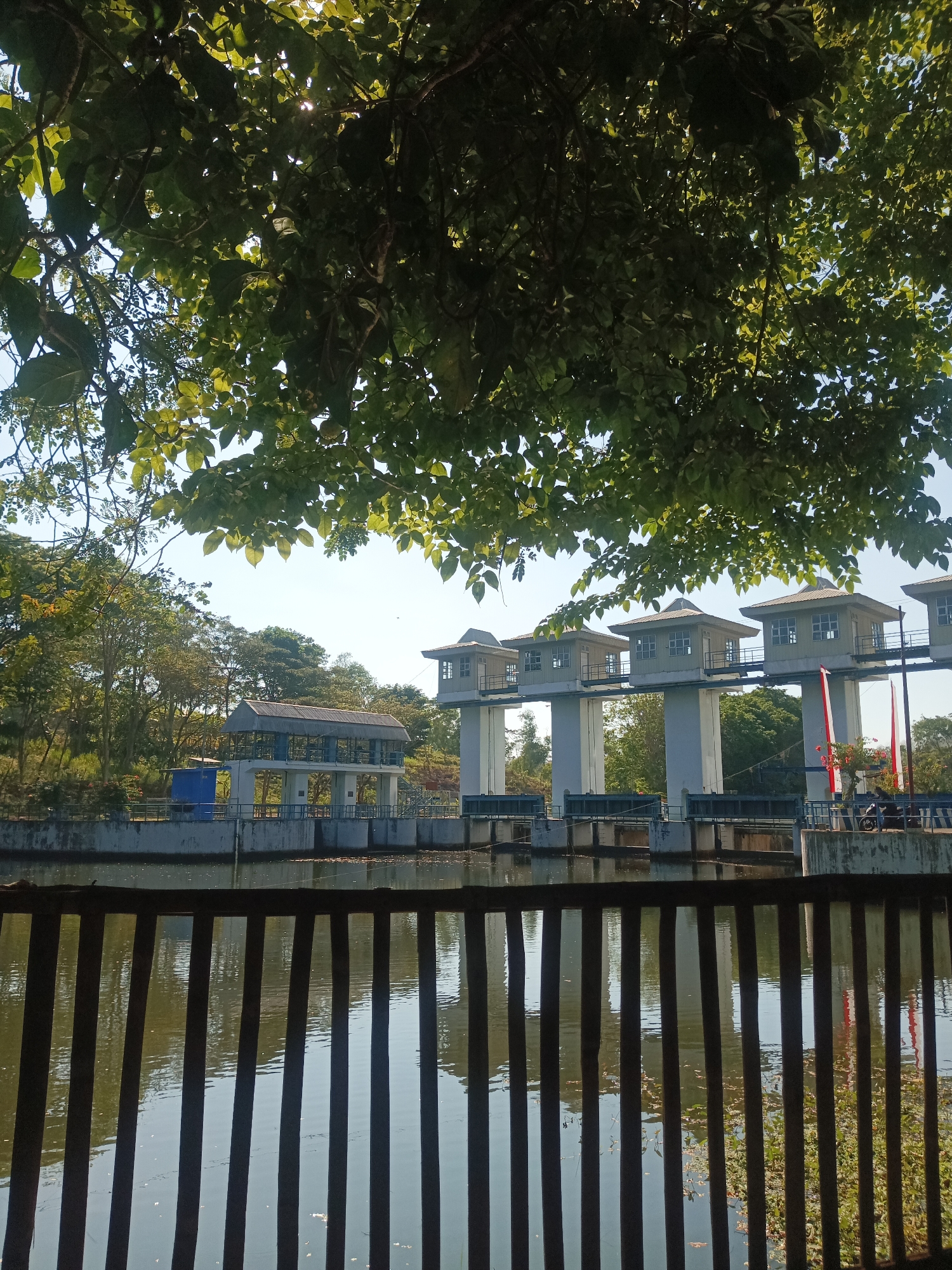 The image depicts a water management structure, likely a sluice gate or dam, situated in the vicinity of Tiudan, Indonesia. The foreground is dominated by a dark, slatted fence that partially obscures a body of water, reflecting the sky and foliage. Beyond the fence and water, the main structure is visible, characterized by several elevated, house-like control booths with windows, supported by tall pillars. Below these booths are water gates and a concrete embankment. To the left of the main structure, a smaller, covered building with blue walls is present. Trees and lush vegetation surround the water management system, with a clear sky overhead, suggesting a daytime scene. The overall impression is one of a functional infrastructure within a natural, green environment.