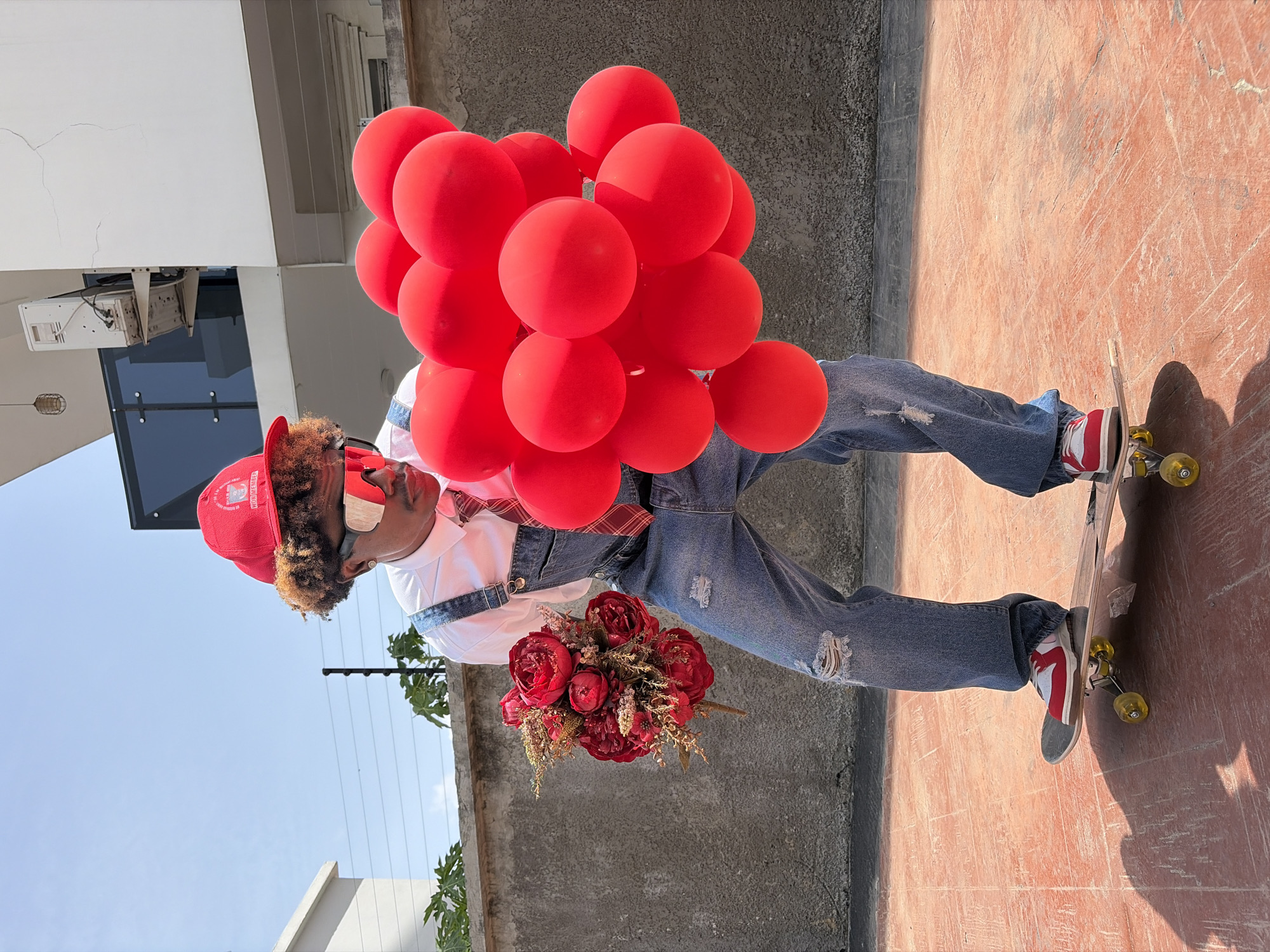 A person, presumably male, stands on a skateboard holding red balloons and a bouquet of red flowers. He wears a red cap, sunglasses, a white t-shirt, denim overalls, and red and white sneakers. His jeans have a distressed, ripped appearance. The background consists of a low concrete wall and a building with a window and air conditioning unit. The setting appears to be outdoors, on a reddish-brown patterned concrete surface. The location is in the city of Igboefon, Nigeria.