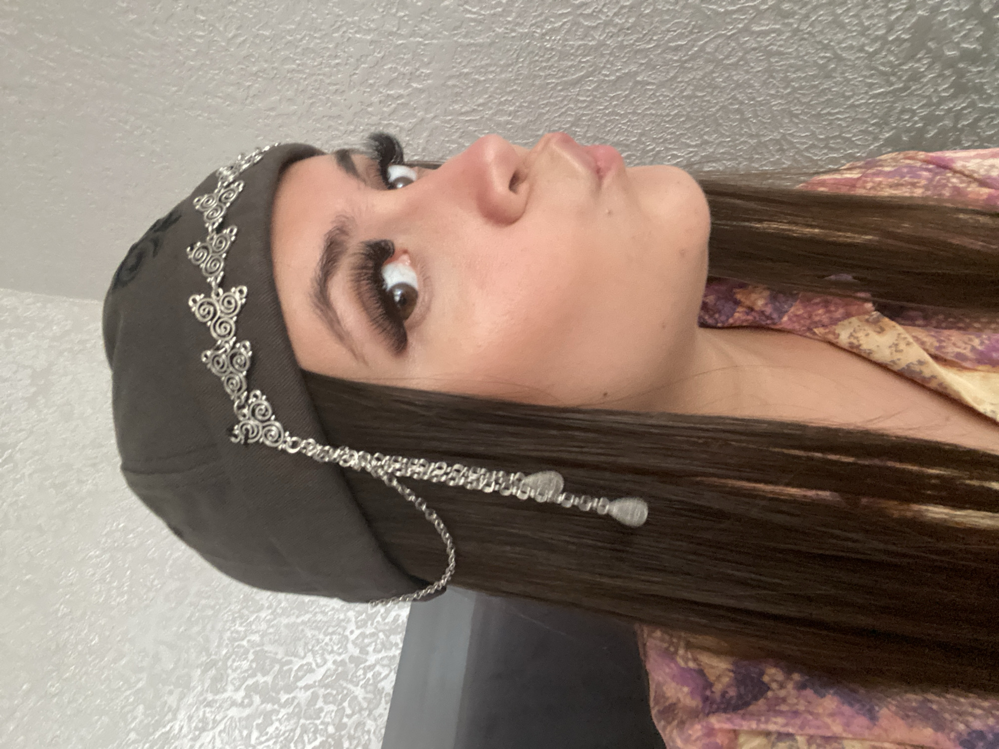 A young woman with dark hair is looking to her right and puckering her lips. She is wearing a dark-colored beret adorned with silver metal scrollwork and hanging chains with teardrop-shaped charms. Her makeup includes long, false eyelashes and winged eyeliner. She is wearing a colorful shirt with a pattern of purple and yellow shapes. The background appears to be a textured gray wall. There is no indication of the location being Chalda, Russia.