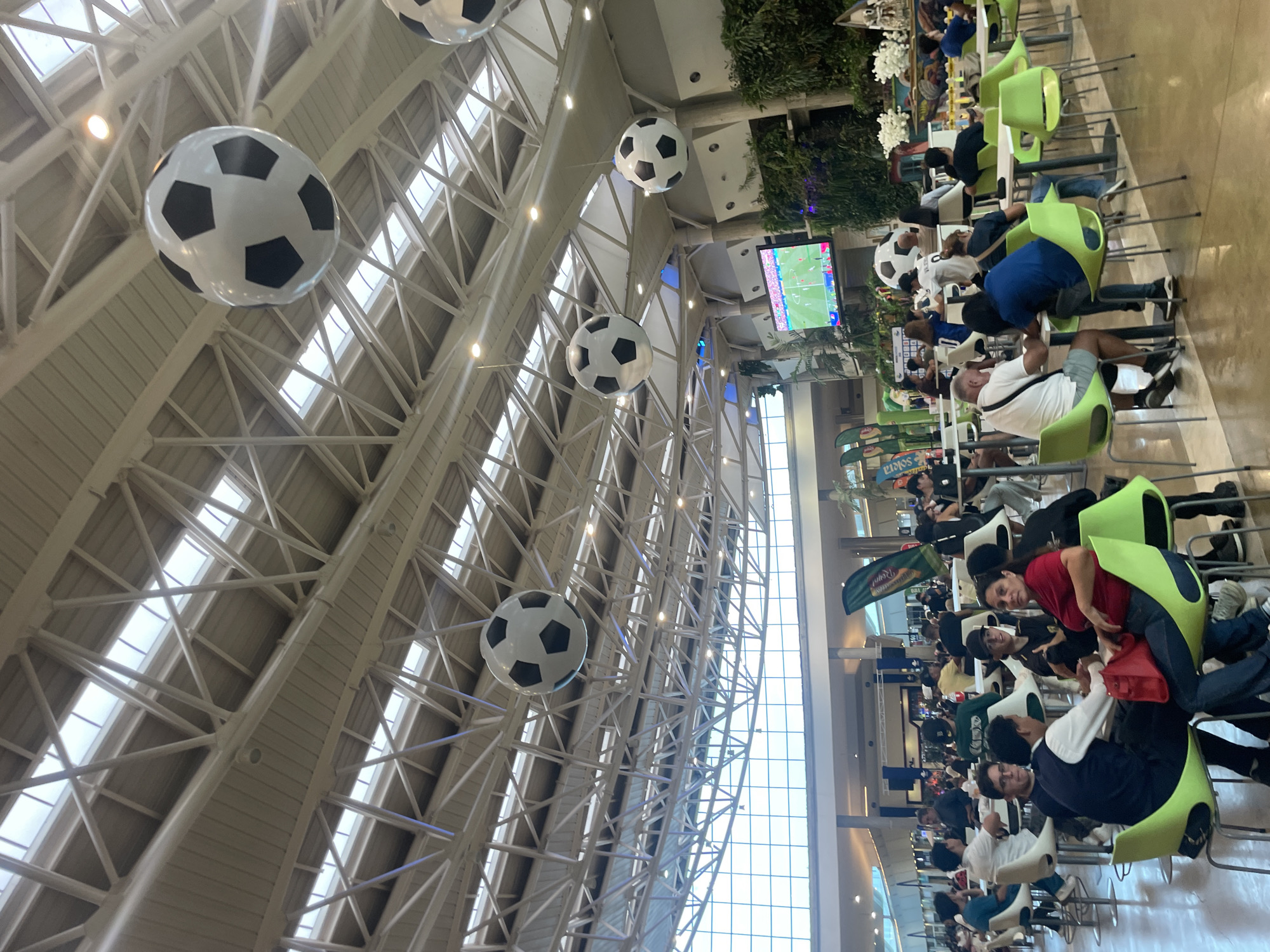 The image depicts a bustling food court area, likely at a shopping mall, in Pampatar, Venezuela. The space is large and open, with a high, exposed ceiling featuring intricate white metal structural supports and numerous skylights that allow natural light to filter in. Several large, inflatable soccer balls are suspended from the ceiling, adding a decorative and playful element to the environment.
Below the ceiling, a large flat-screen television is mounted high on a wall, displaying a soccer match. The food court is filled with people seated at tables, engaged in conversation, eating, and watching the television. The seating consists of modern, brightly colored chairs, primarily lime green, arranged around white tables. The flooring is a polished, light-colored material.
Various vendors' stalls are visible in the background, some with awnings and signage, suggesting a commercial setting. There are also lush green plants and floral arrangements, adding to the ambiance. The overall scene is one of a lively public gathering space, with a clear focus on a sporting event, indicated by the soccer balls and the televised match. The time of day appears to be daytime, given the presence of natural light from the skylights.